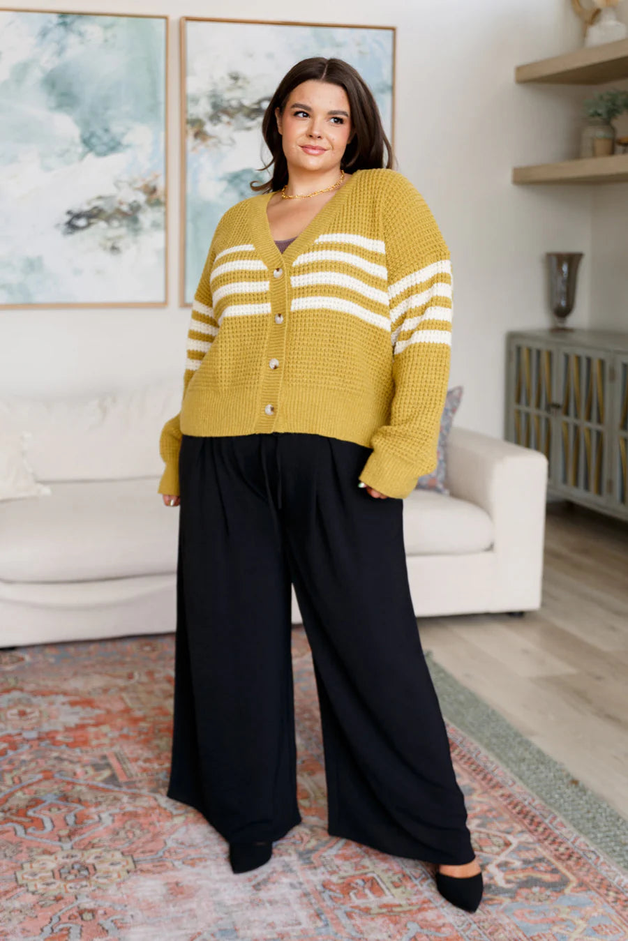 Plus-size woman wearing mustard knit cardigan with white stripes and black wide-leg pants indoors in stylish room