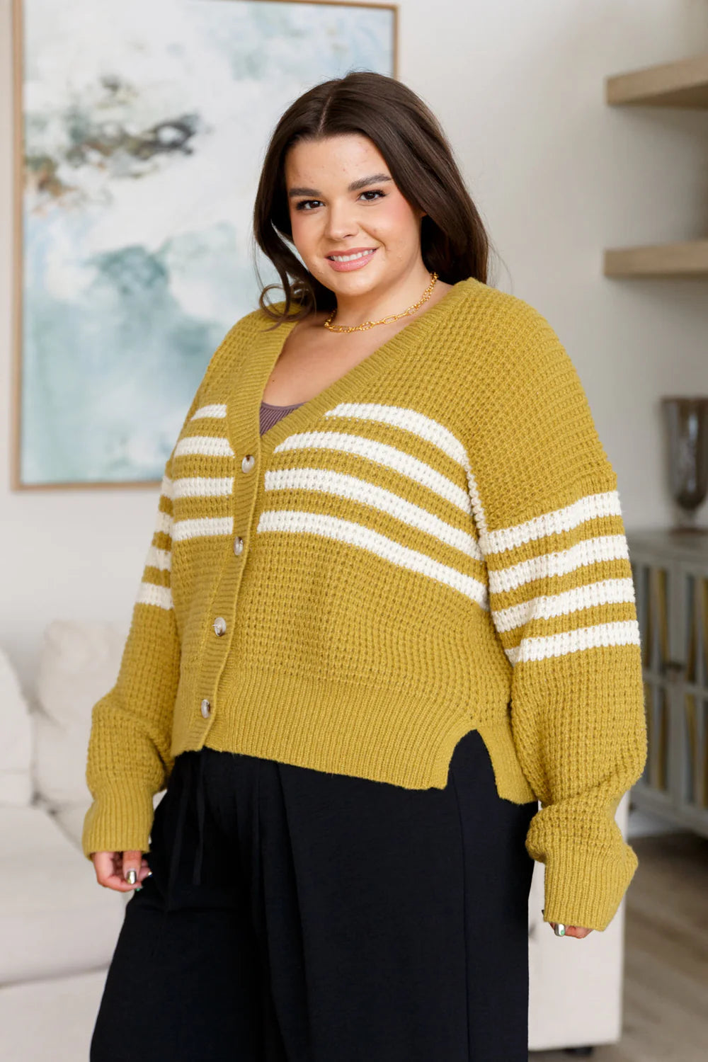 Smiling woman wearing mustard knit cardigan with white stripes and black wide-leg pants indoors in cozy room