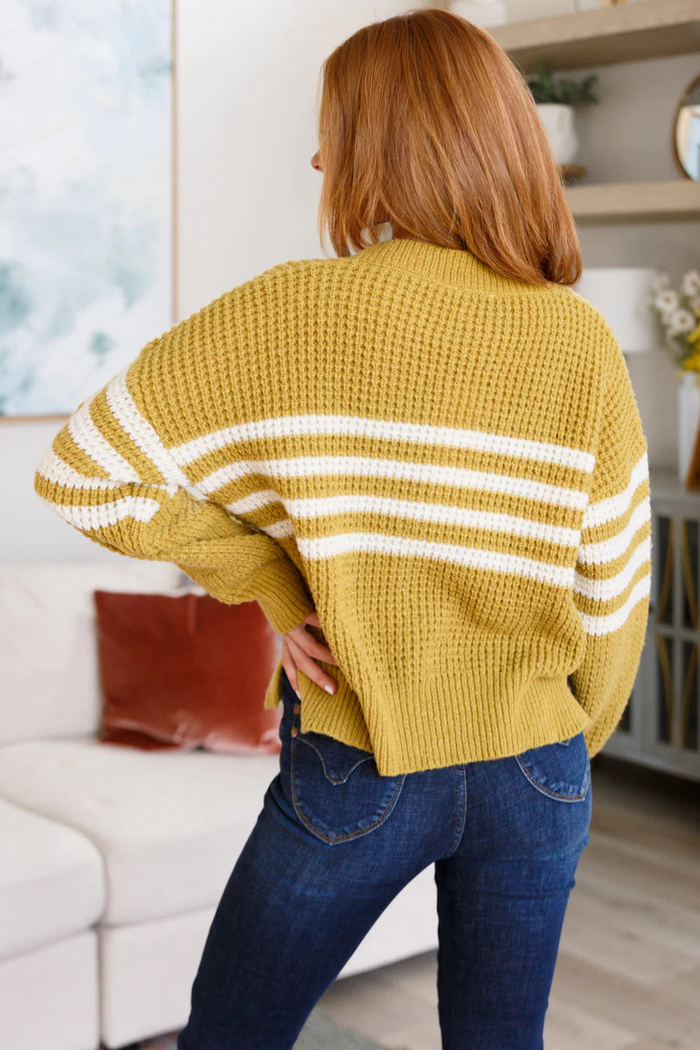 Woman with red hair wearing mustard striped knit cardigan and dark jeans standing indoors with hands on hips