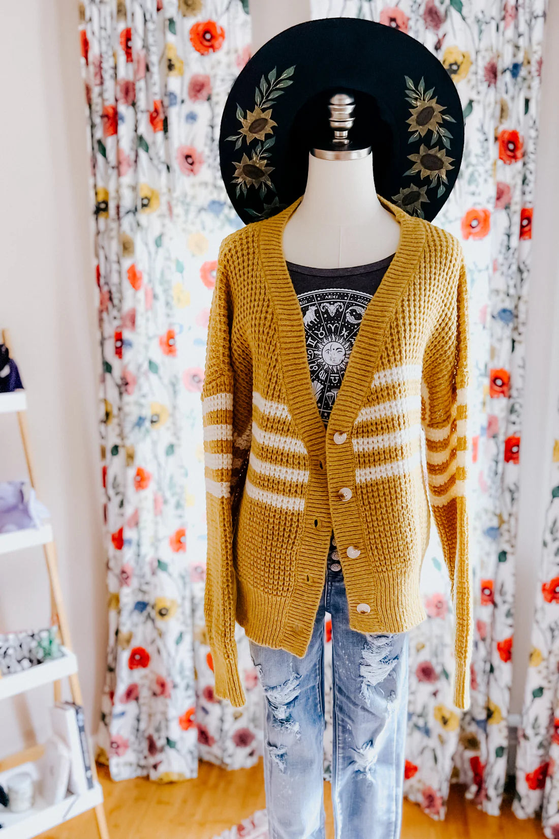 Mustard knit cardigan with white stripes layered over graphic tee and distressed jeans on mannequin with floral backdrop