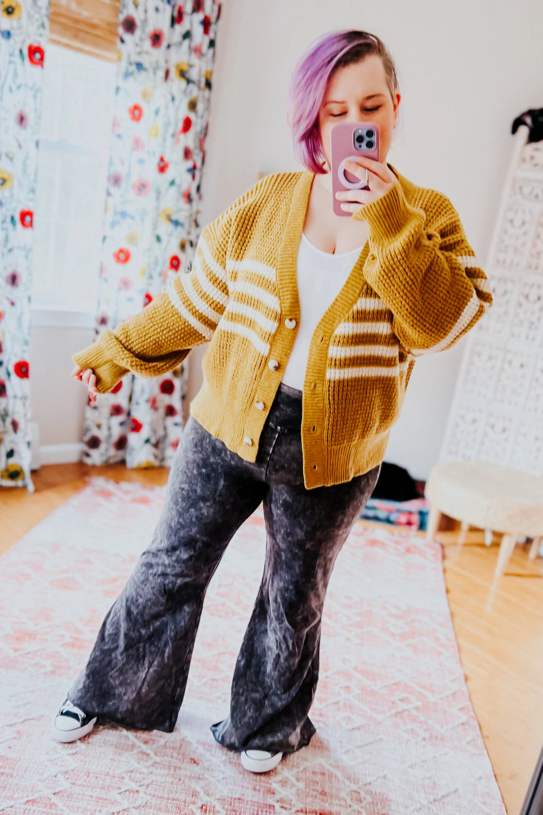 Woman with lavender hair wearing a mustard striped knit cardigan over white top with dark washed flare pants in cozy room