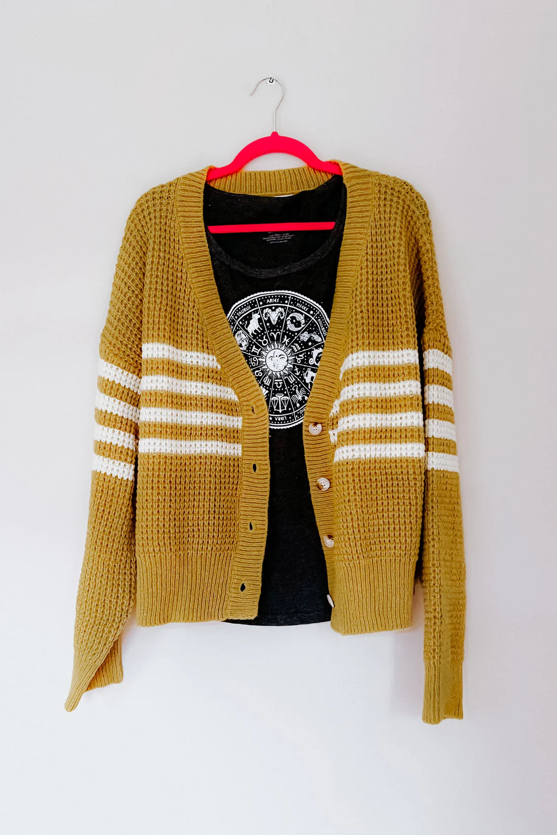 Mustard knit cardigan with white stripes over black graphic top hanging on pink hanger against white wall