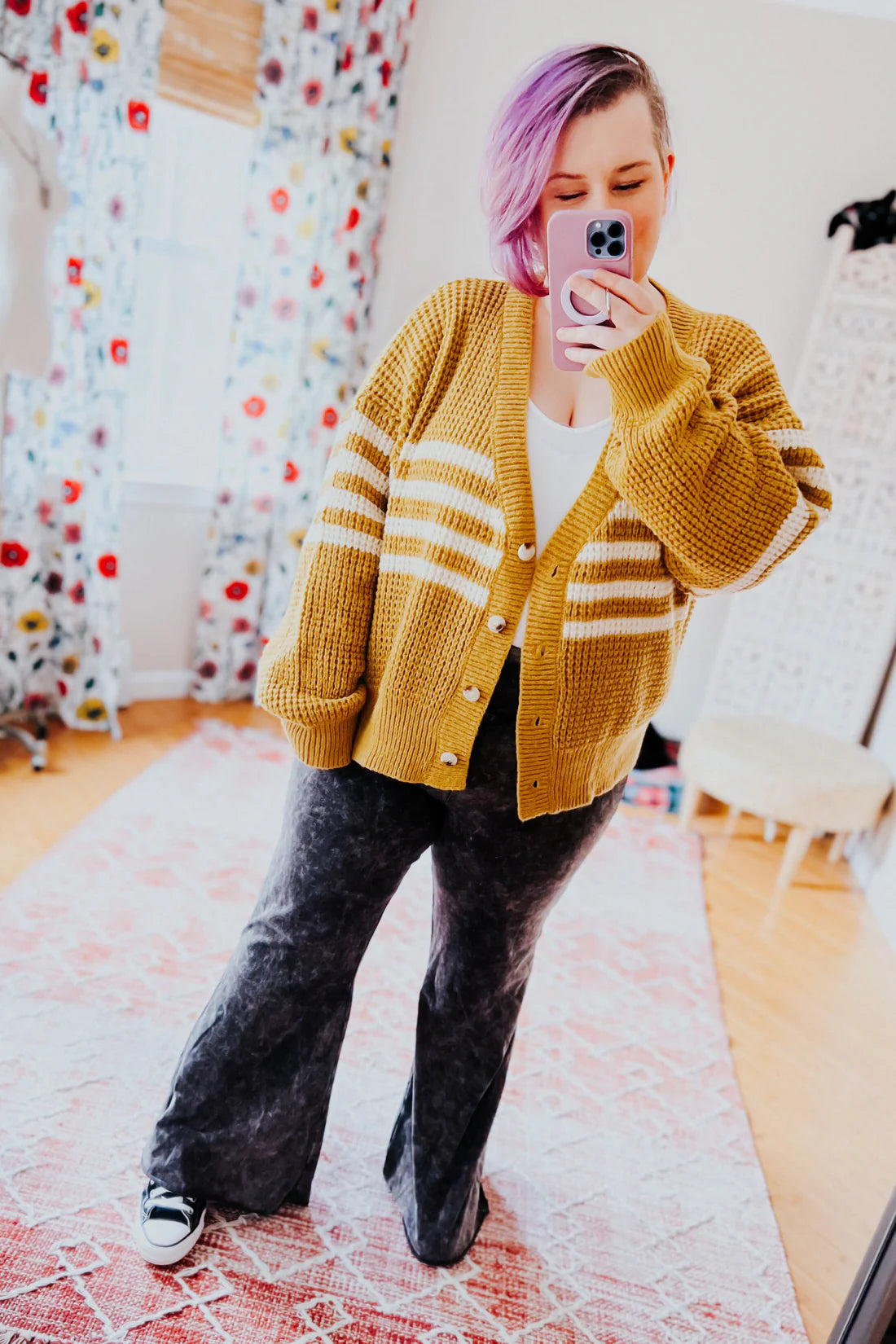 Woman with short lavender hair wearing mustard striped cardigan and black flared pants taking a mirror selfie indoors