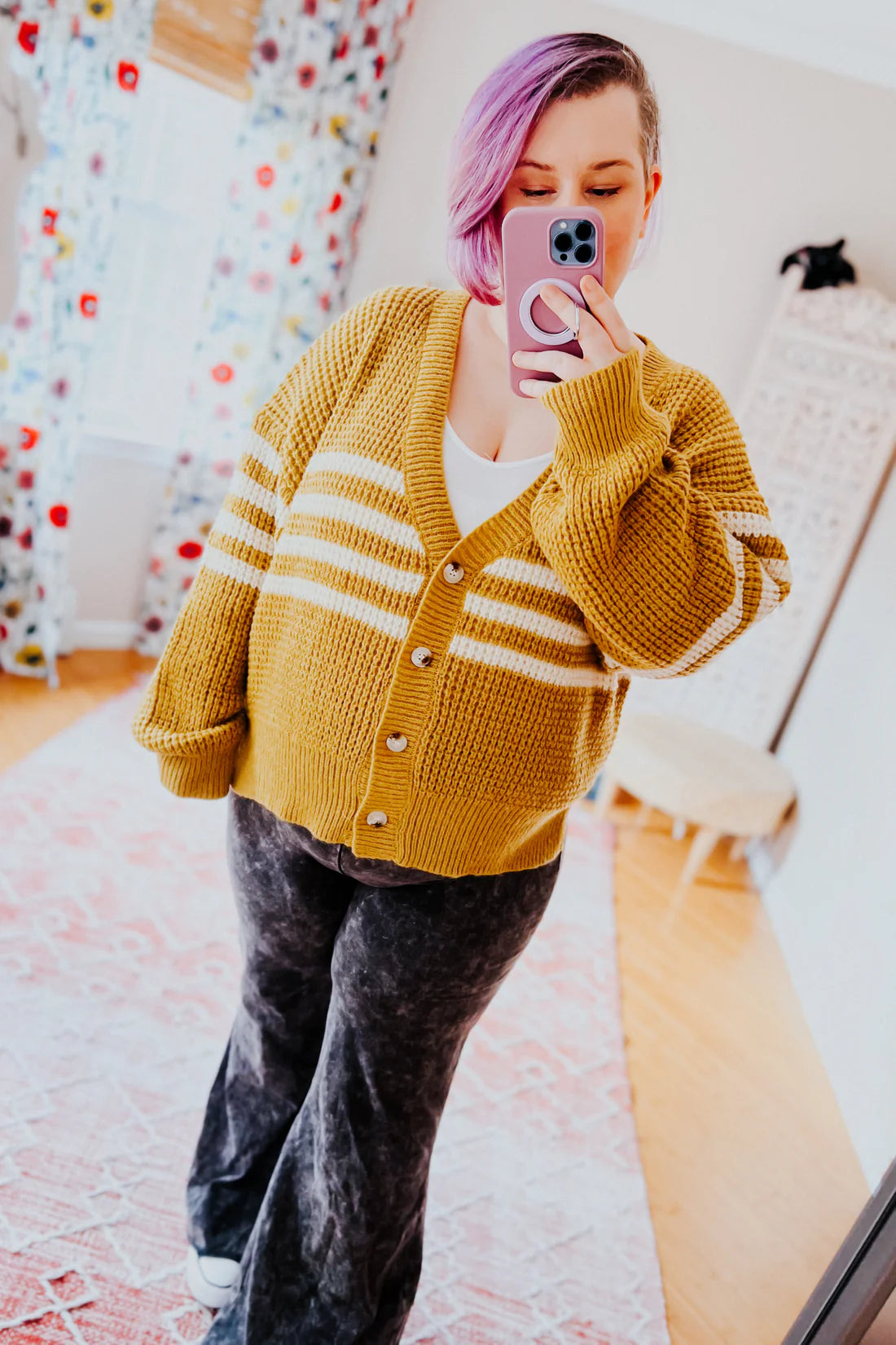 Woman with lavender hair wearing mustard and cream striped cardigan and black pants taking a mirror selfie indoors