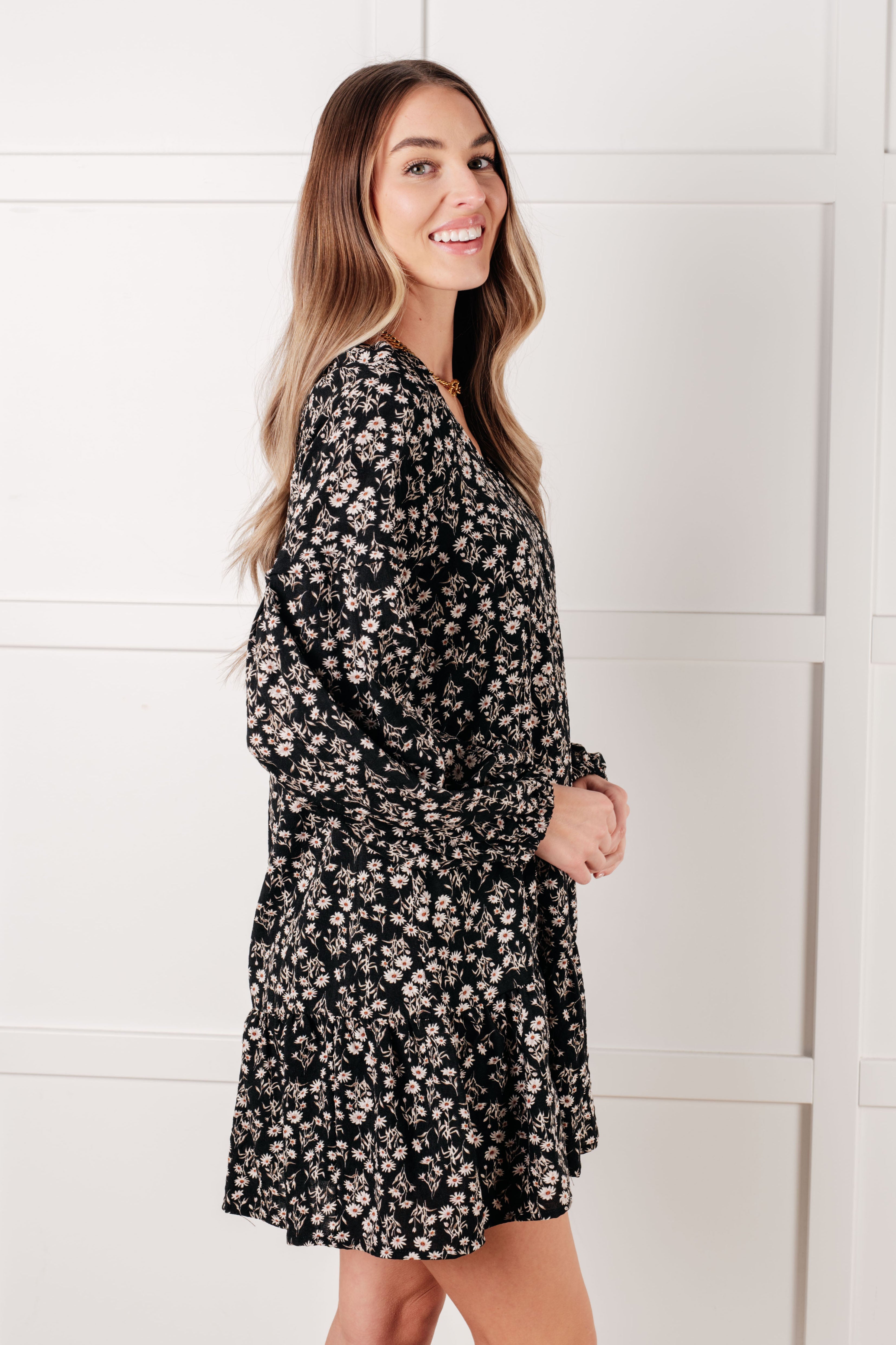 Woman smiling wearing black floral v-neck dress with long gathered sleeves and relaxed fit against white wall
