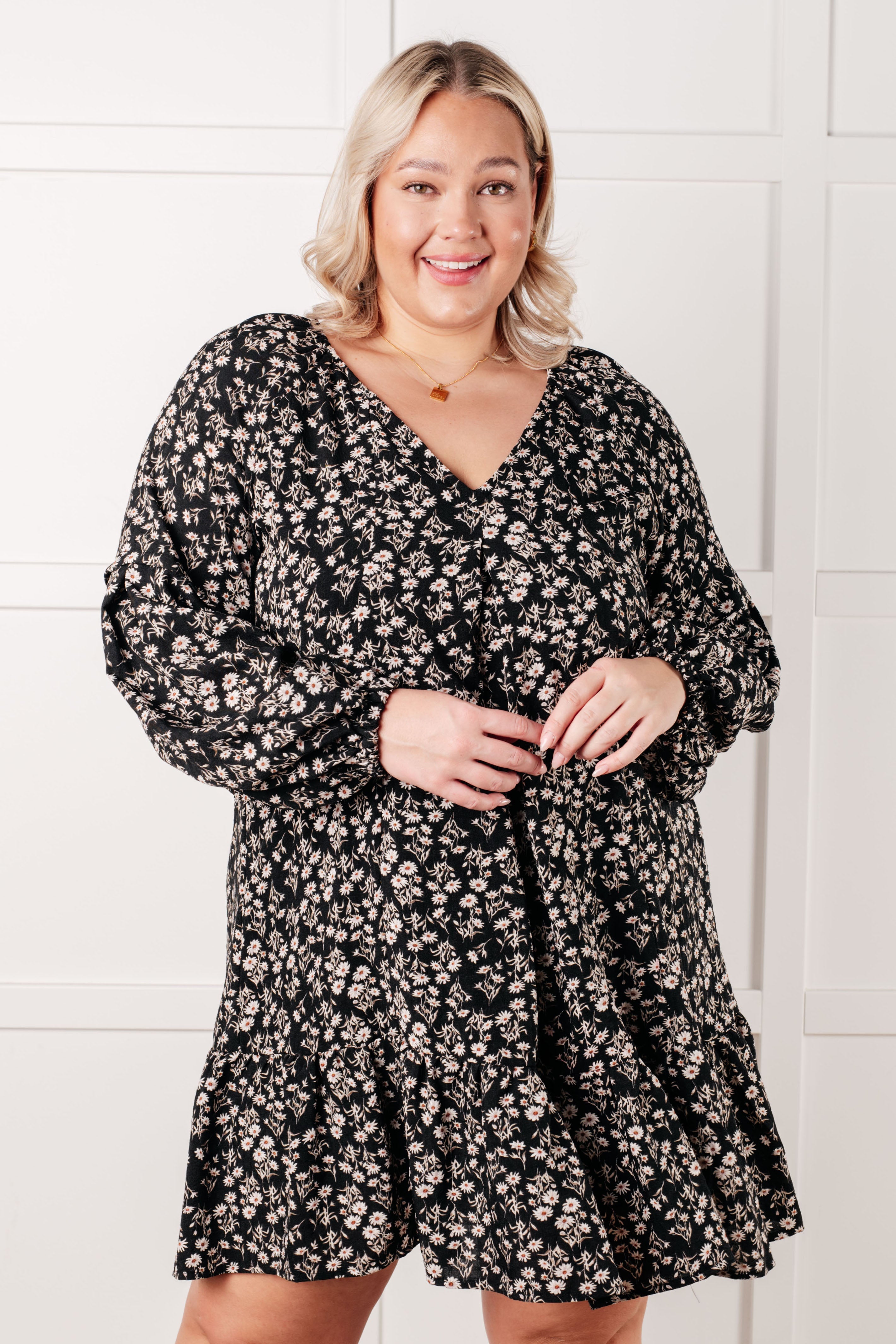 Plus size model wearing black floral v-neck dress with voluminous sleeves and delicate pendant necklace indoors