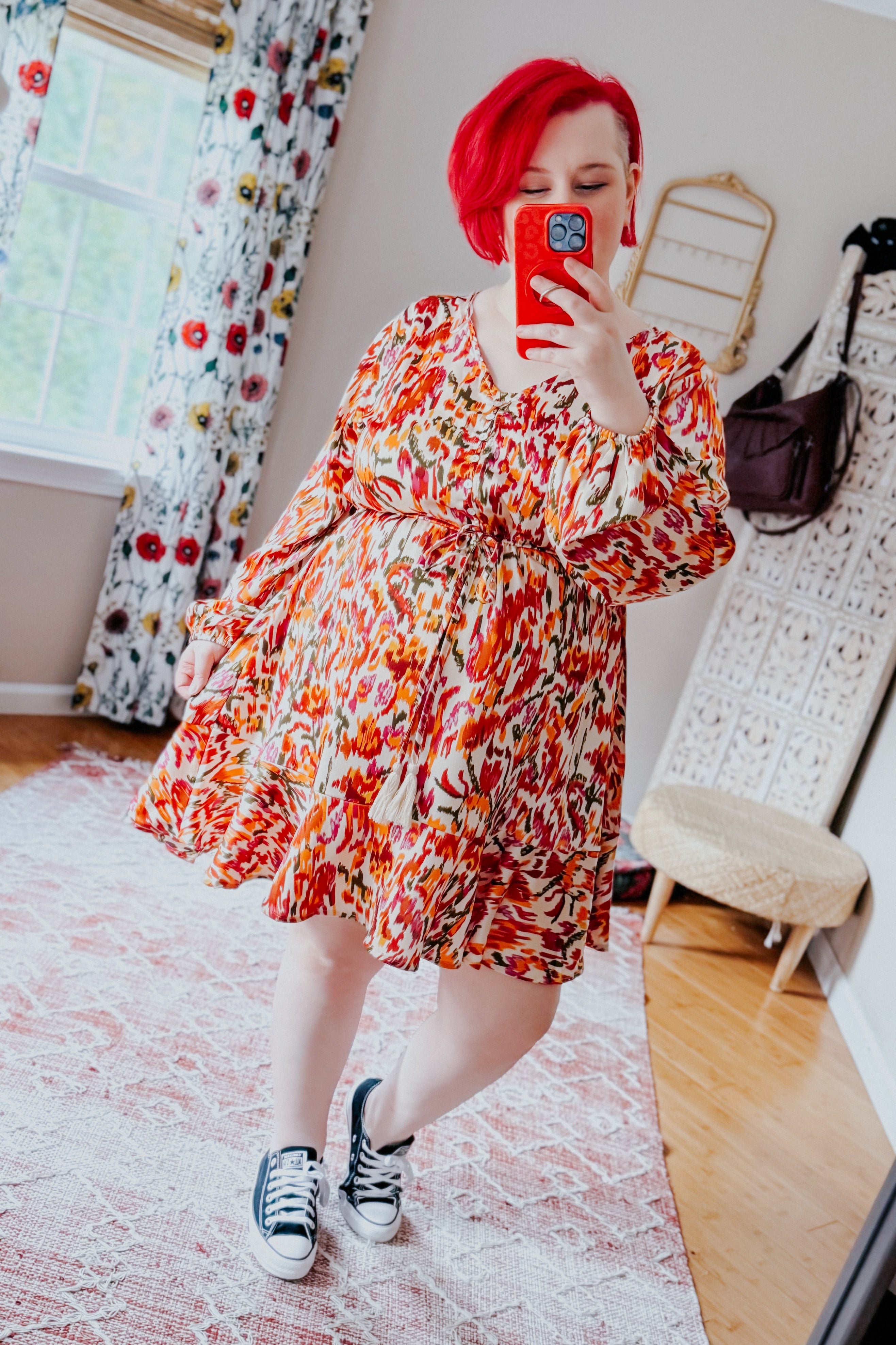 Plus size woman with red hair wearing orange patterned V-neck dress with balloon sleeves and tie waist, taking mirror selfie indoors