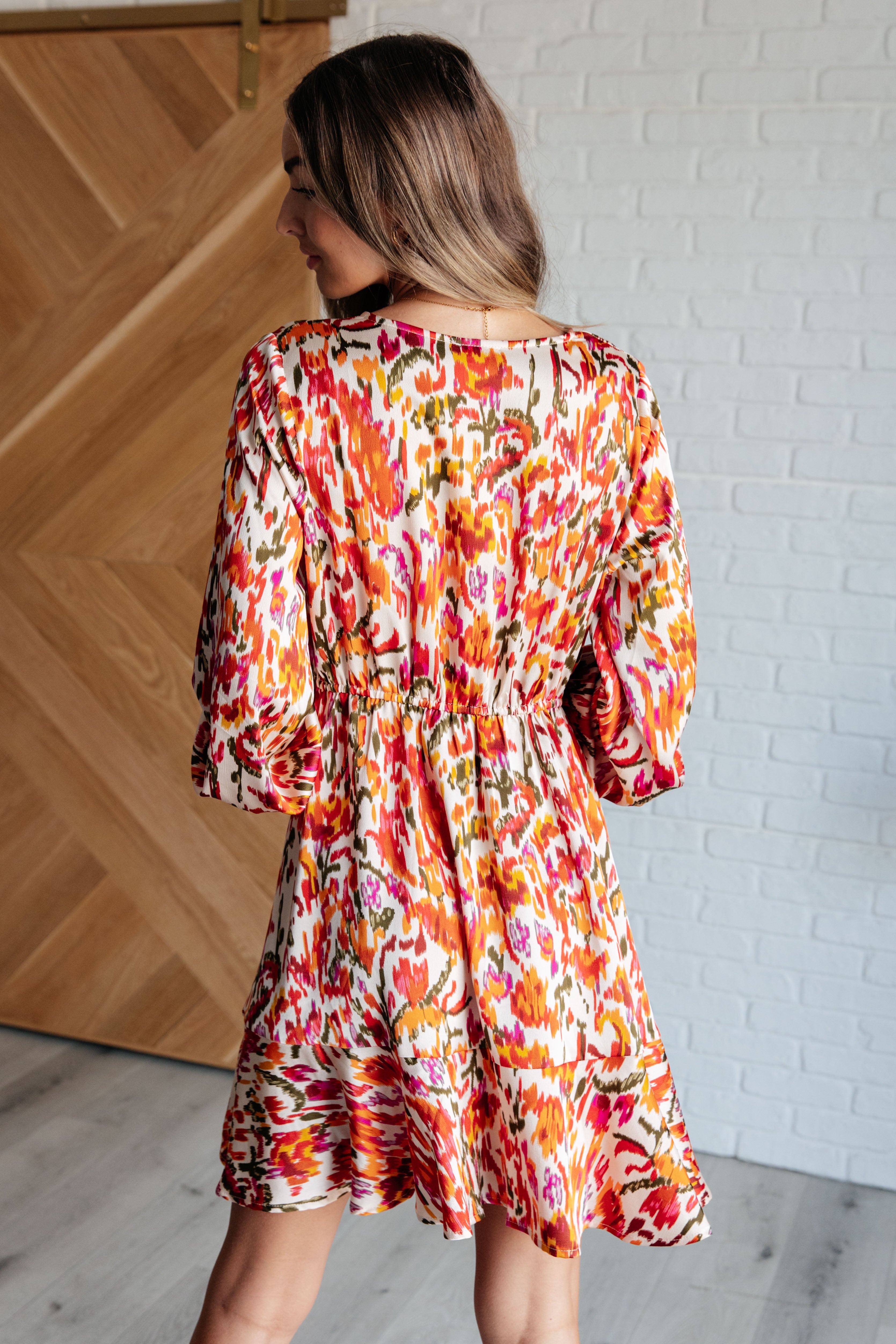 Woman wearing colorful orange patterned dress with balloon sleeves and gathered waist shown from the back indoors