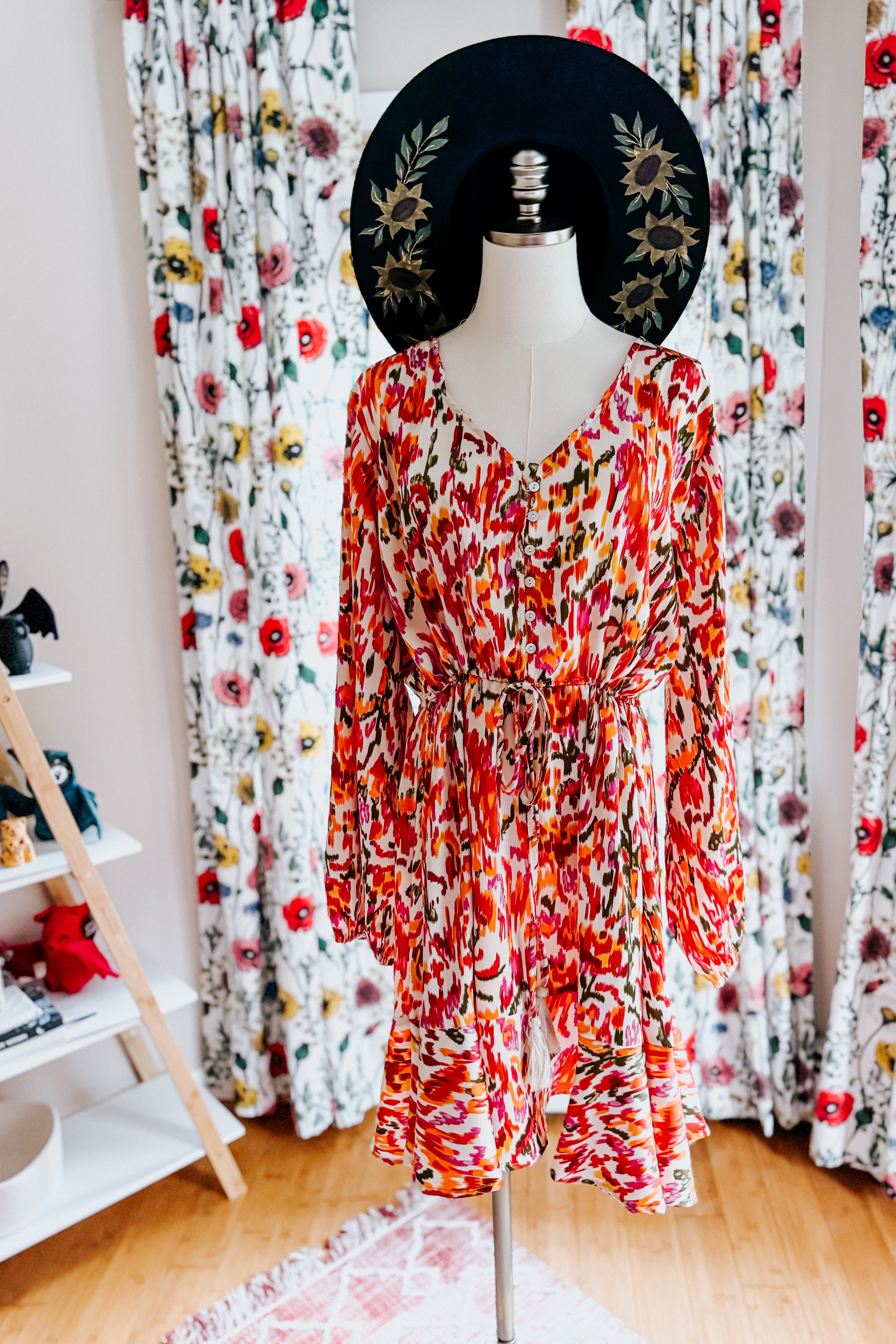 Orange patterned dress with balloon sleeves and tie waist displayed on mannequin with black floral hat indoors