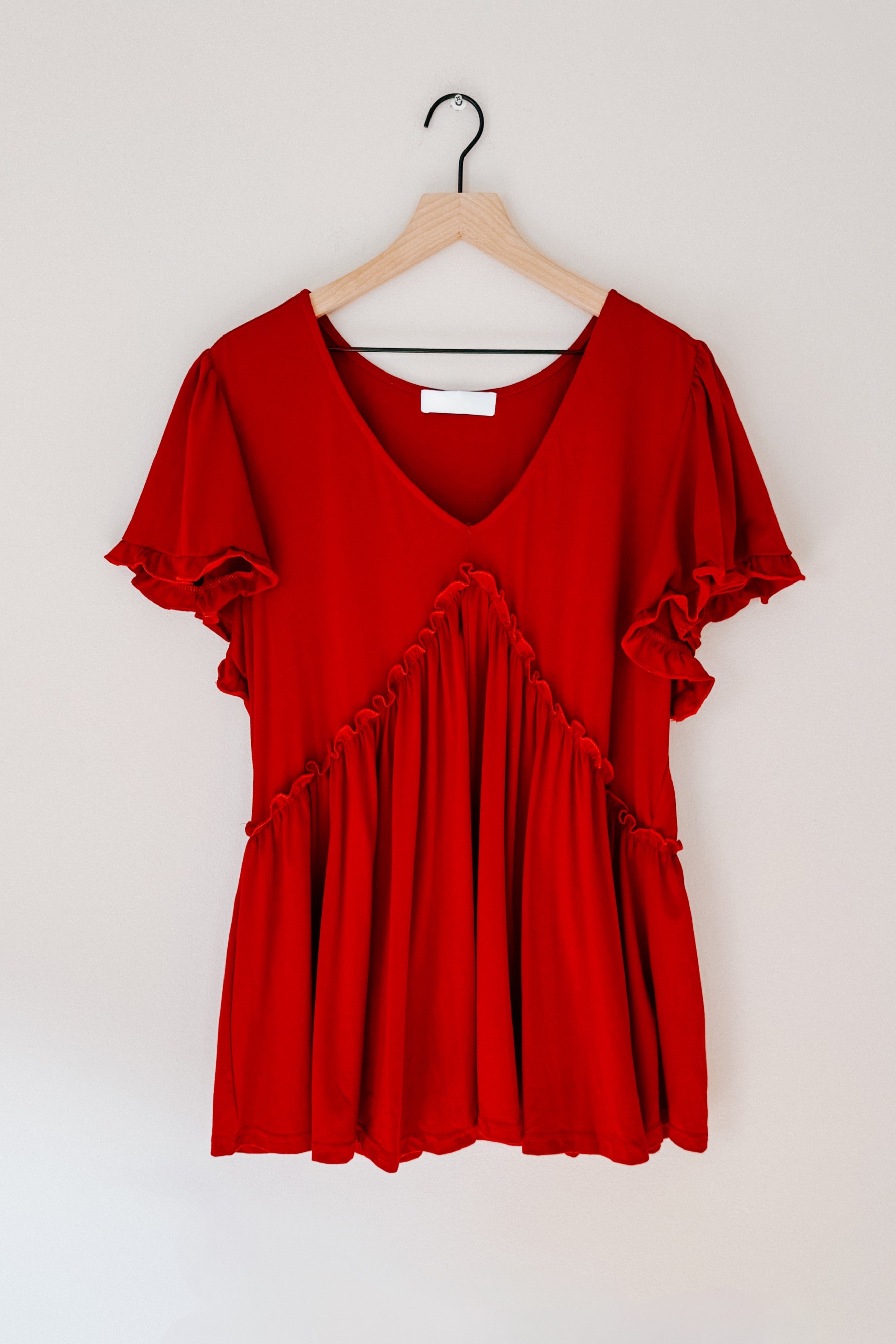Red V-neck top with ruffled diagonal detailing and flutter sleeves hanging on wooden hanger against neutral wall