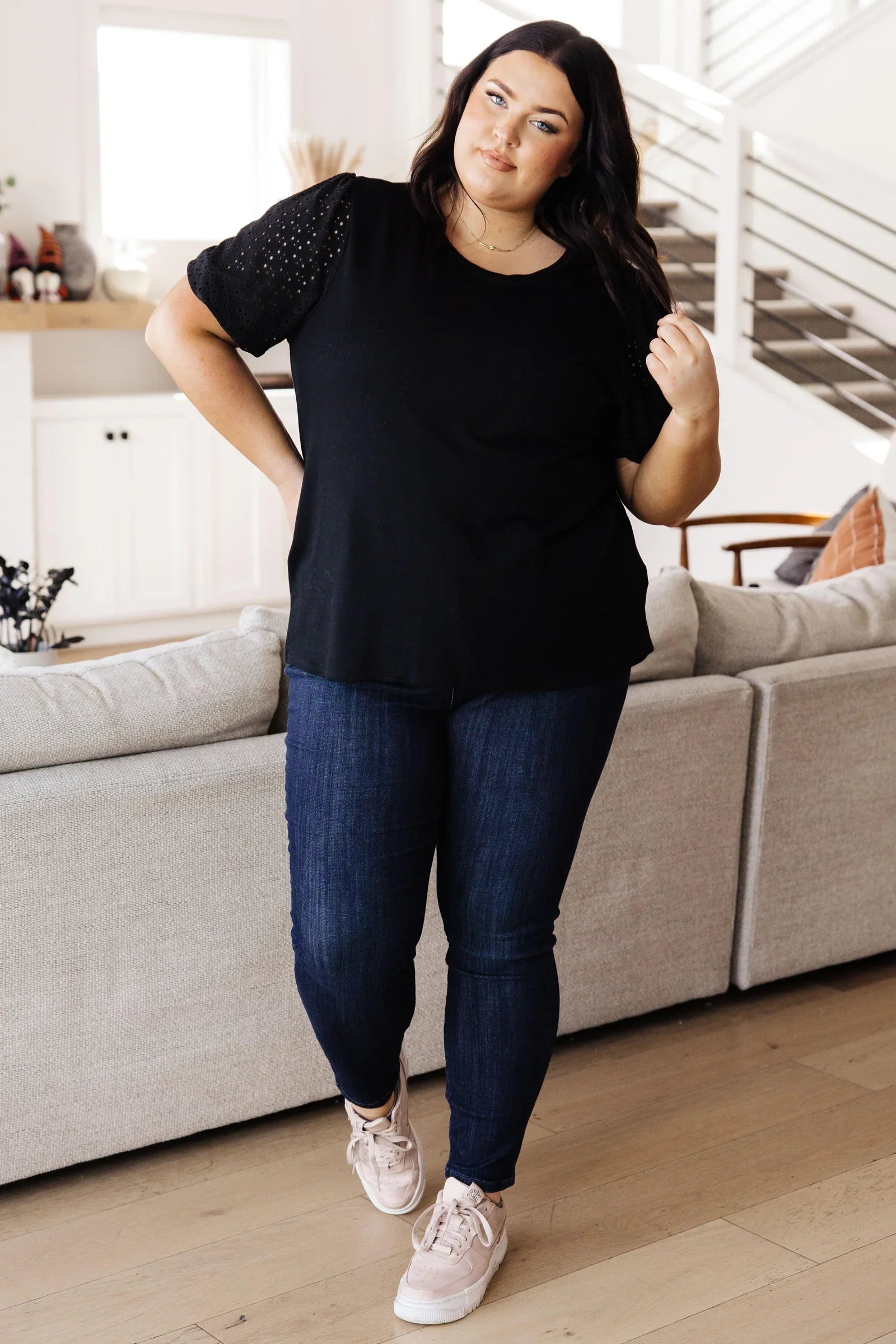 Curvy woman with dark hair wearing a black lace sleeve top and dark blue jeans standing in a bright room