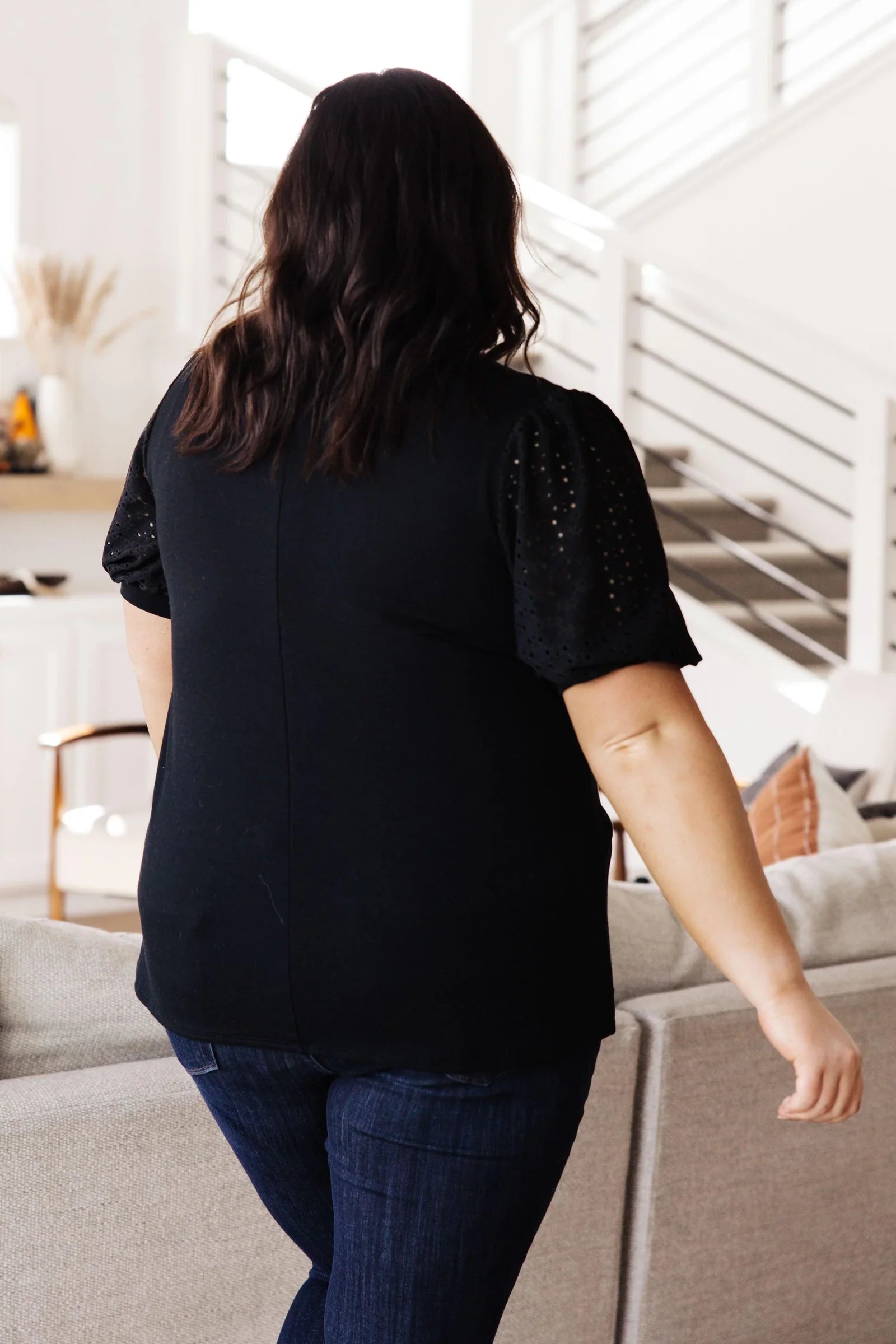 Plus size woman seen from behind wearing a black top with lace puff sleeves and dark blue jeans in a bright modern space