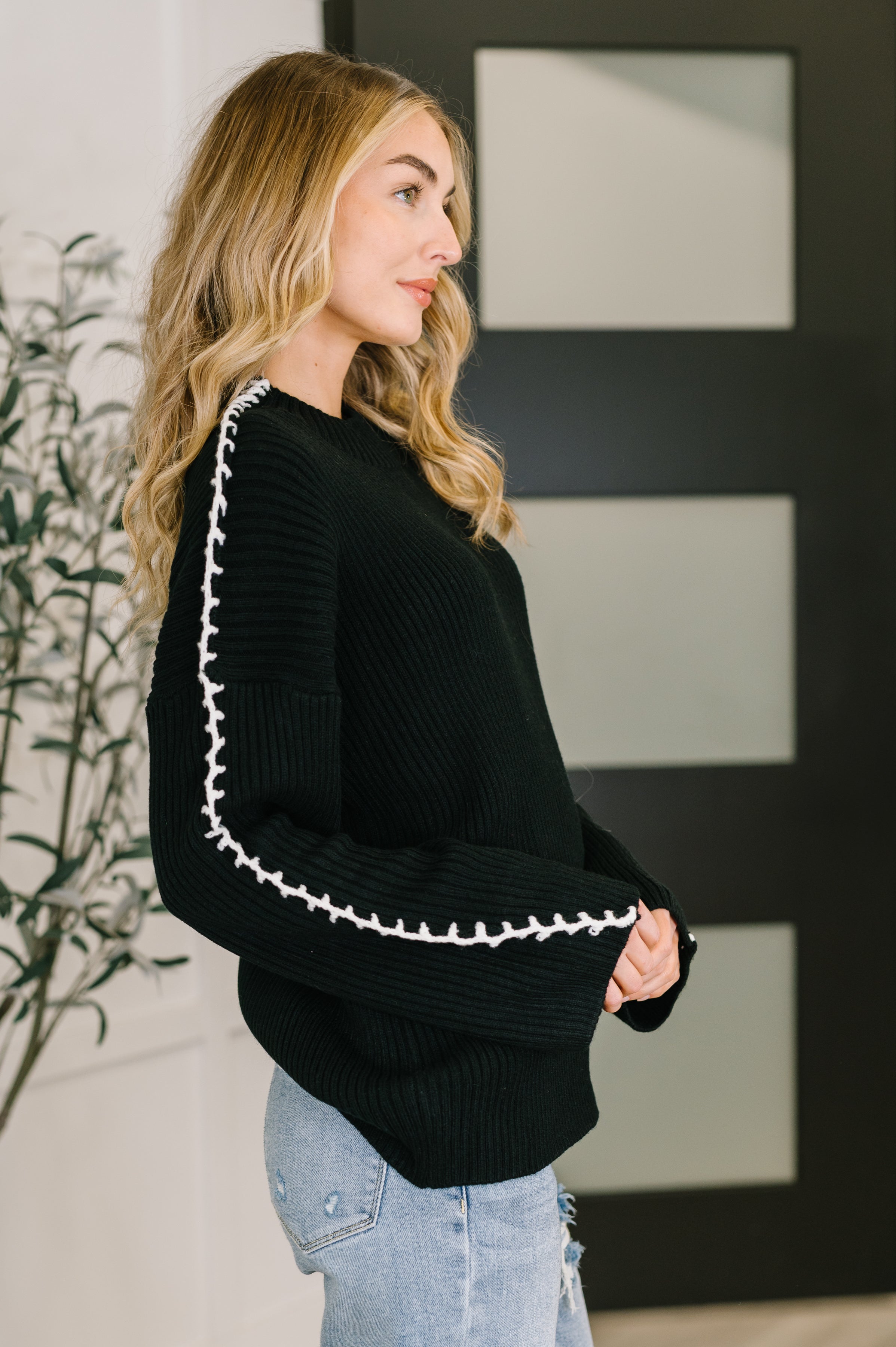 Woman wearing black knit pullover with white stitch details along sleeves paired with light ripped jeans indoors