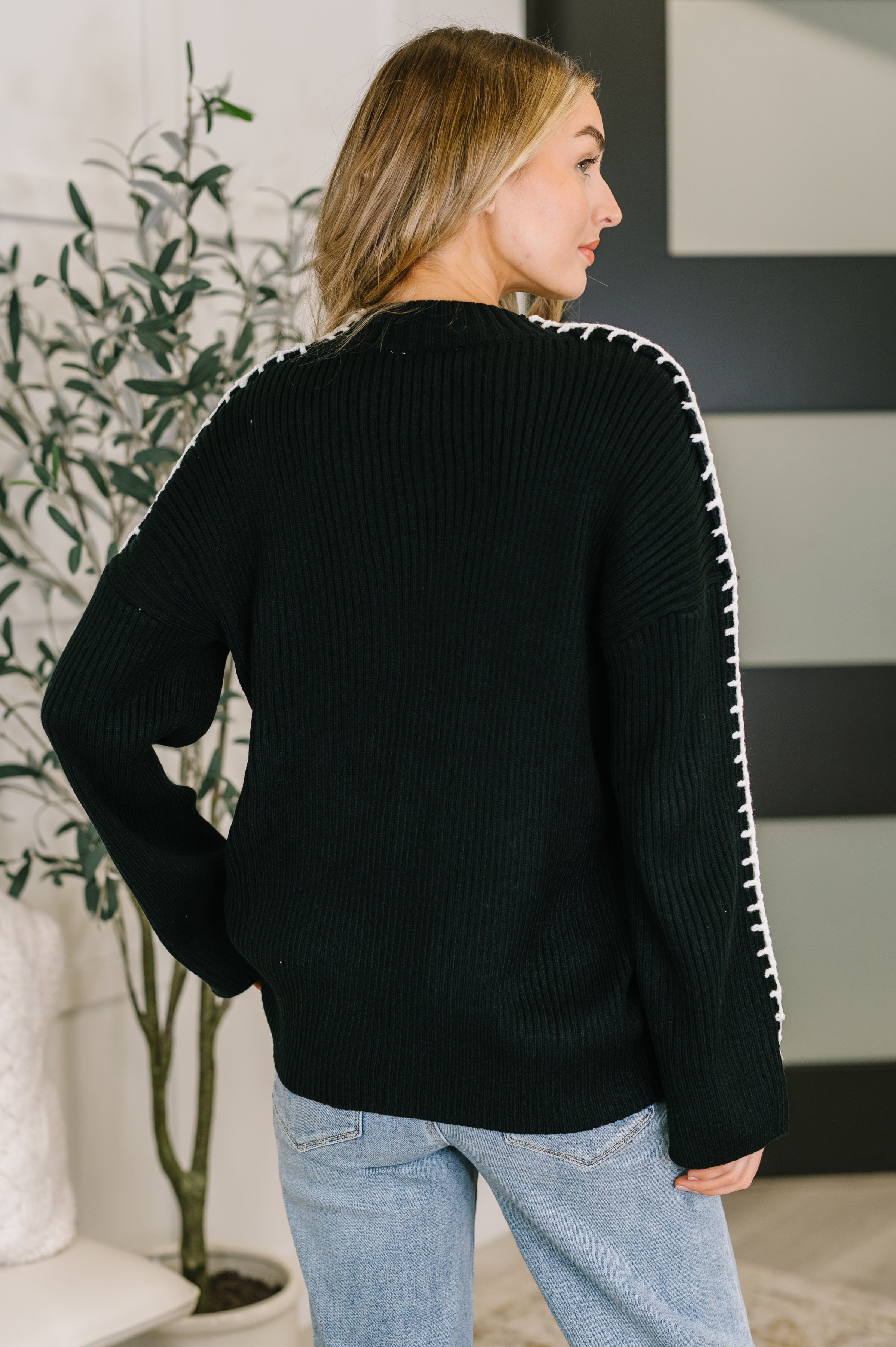 Woman seen from behind wearing a black knit pullover with white stitch details along sleeves paired with light blue jeans indoors