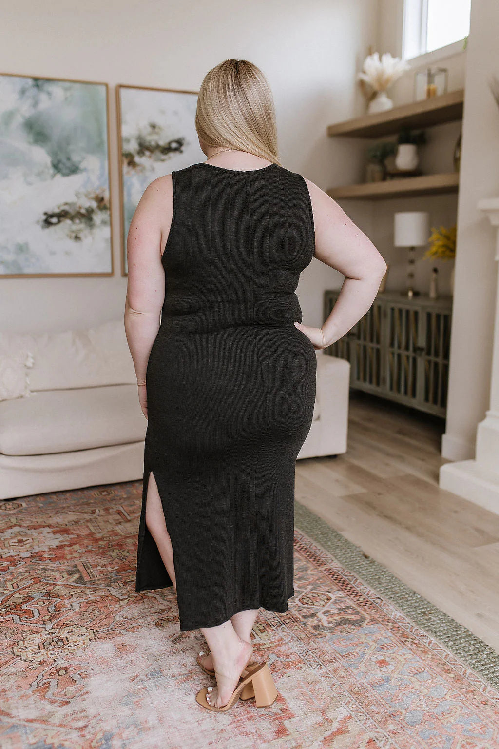 Plus-size woman wearing a sleeveless charcoal midi dress with side slit and block heels in a modern living room