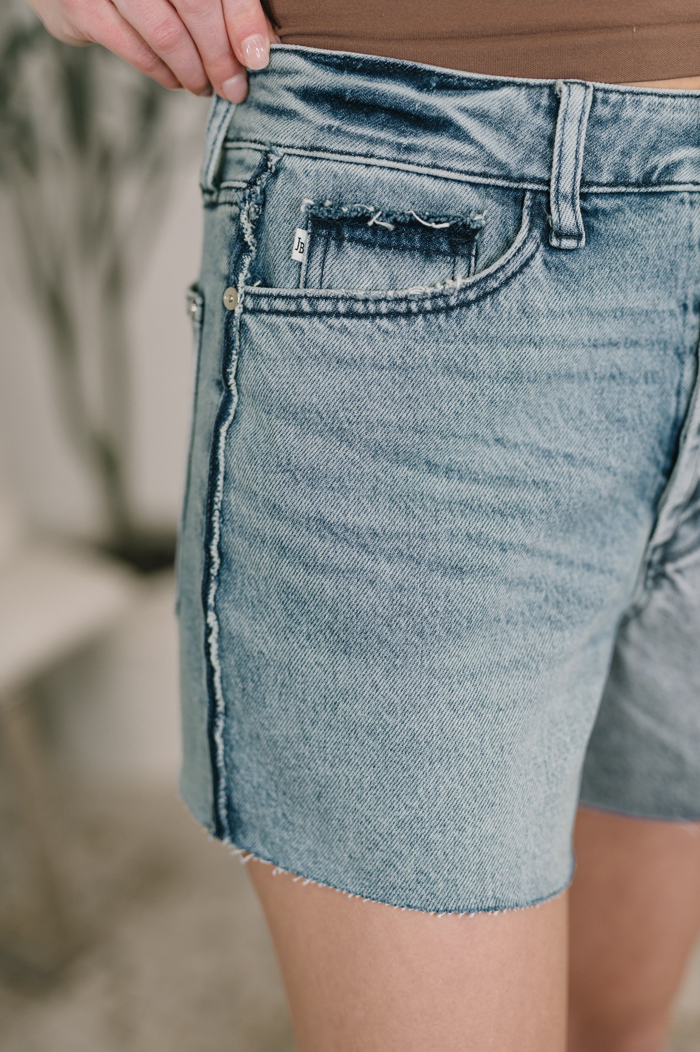 Close-up of blue Paige High Waist Rigid Magic Cutoff Shorts featuring raw side seams and frayed pocket detail indoors