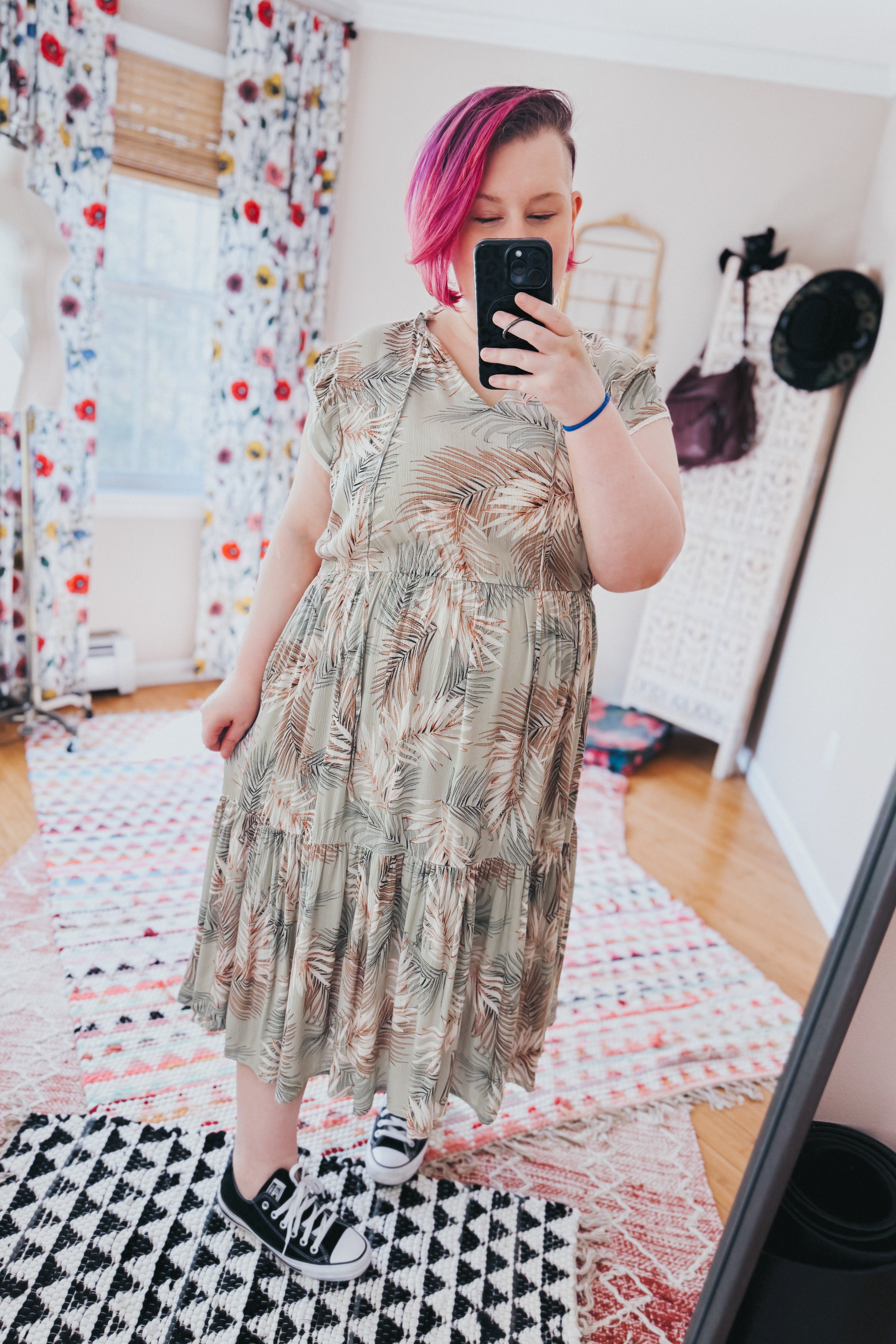Woman with short vibrant pink hair wearing muted green tiered midi dress with palm print and black sneakers indoors
