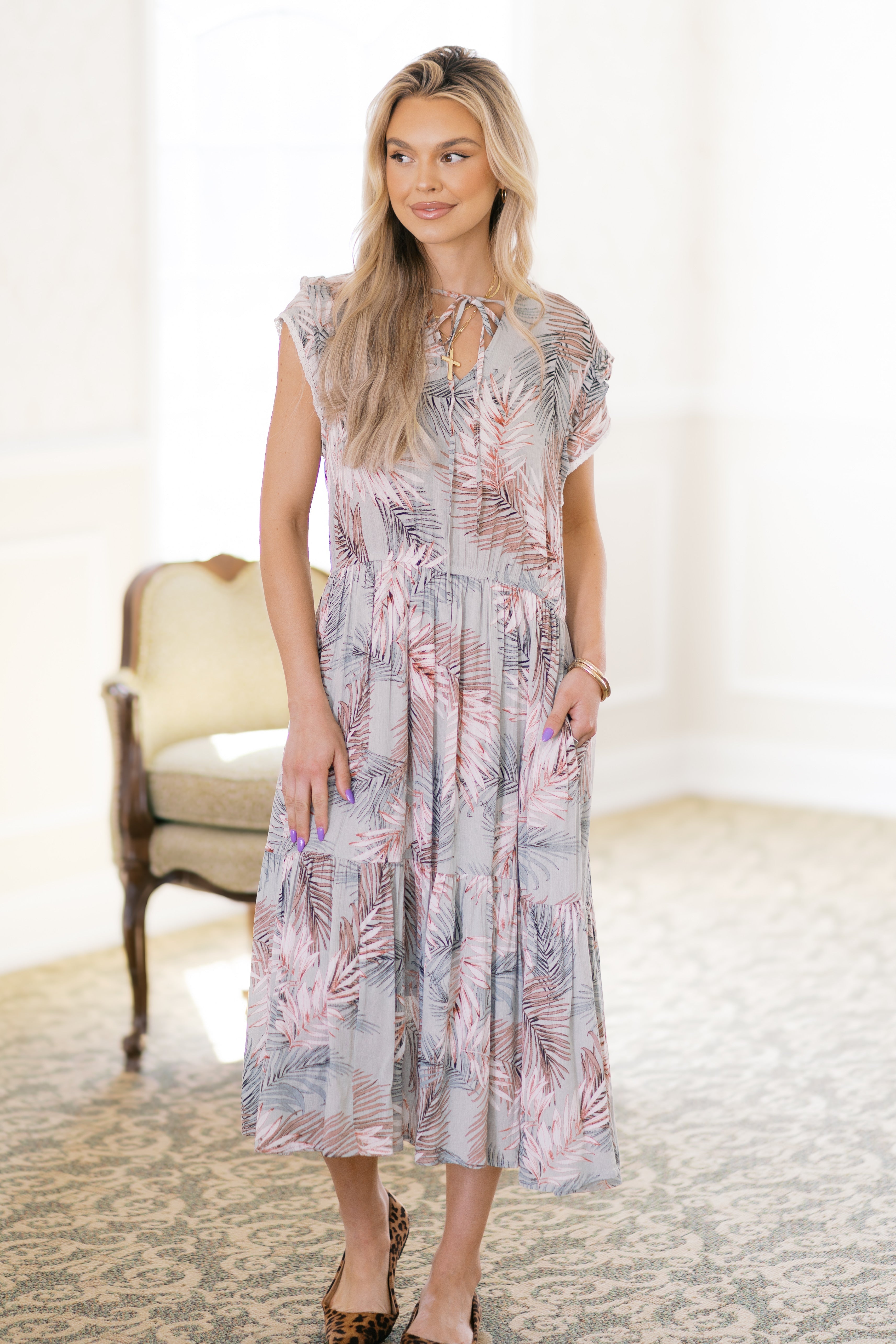 Blonde woman wearing green tiered midi dress with palm leaf pattern and leopard flats standing indoors near vintage chair
