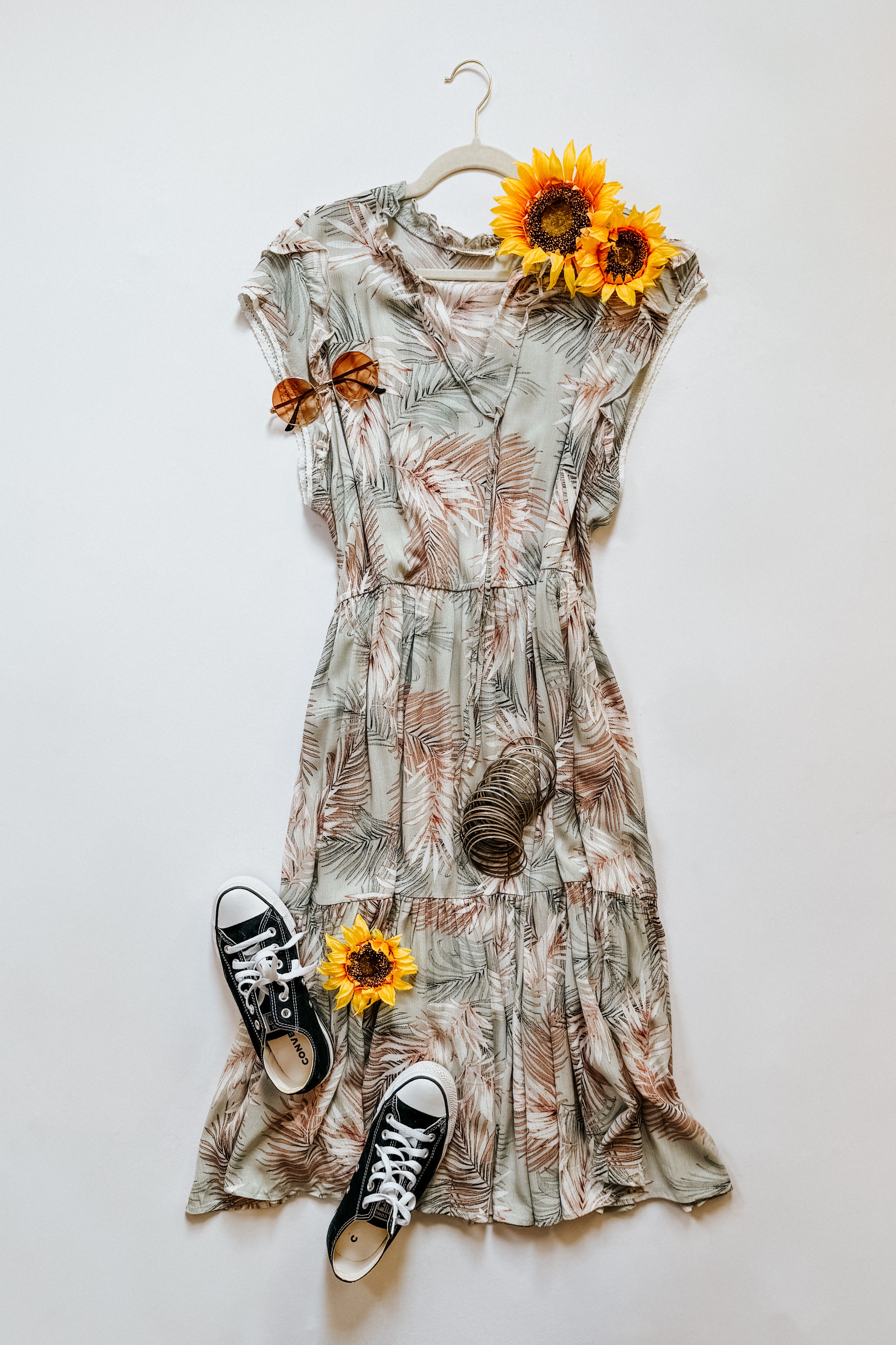 Green tiered midi dress with palm leaf print styled with black sneakers, round sunglasses, and sunflowers on white background