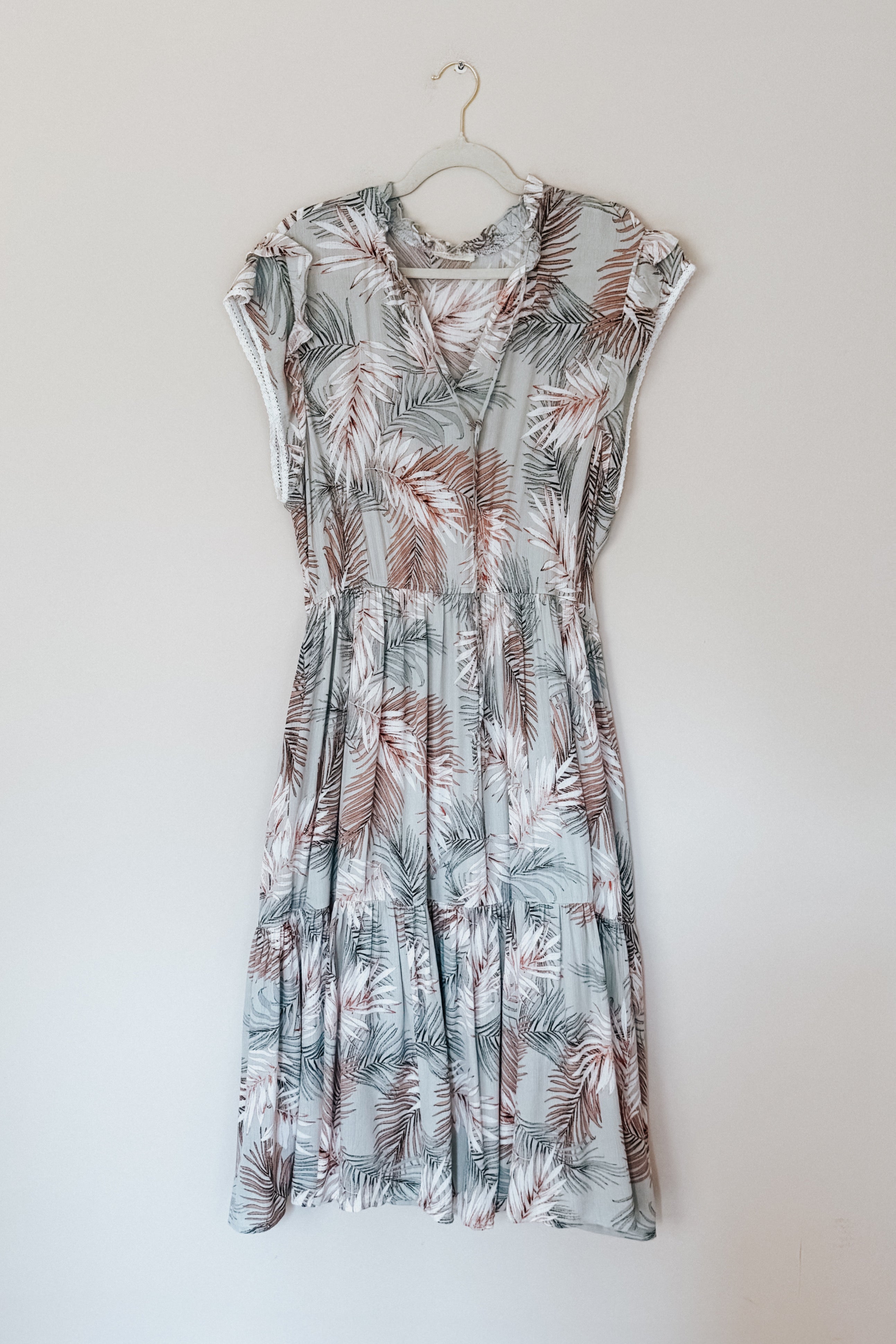 Sleeveless green tiered midi dress with brown and green palm leaf print hanging against plain wall