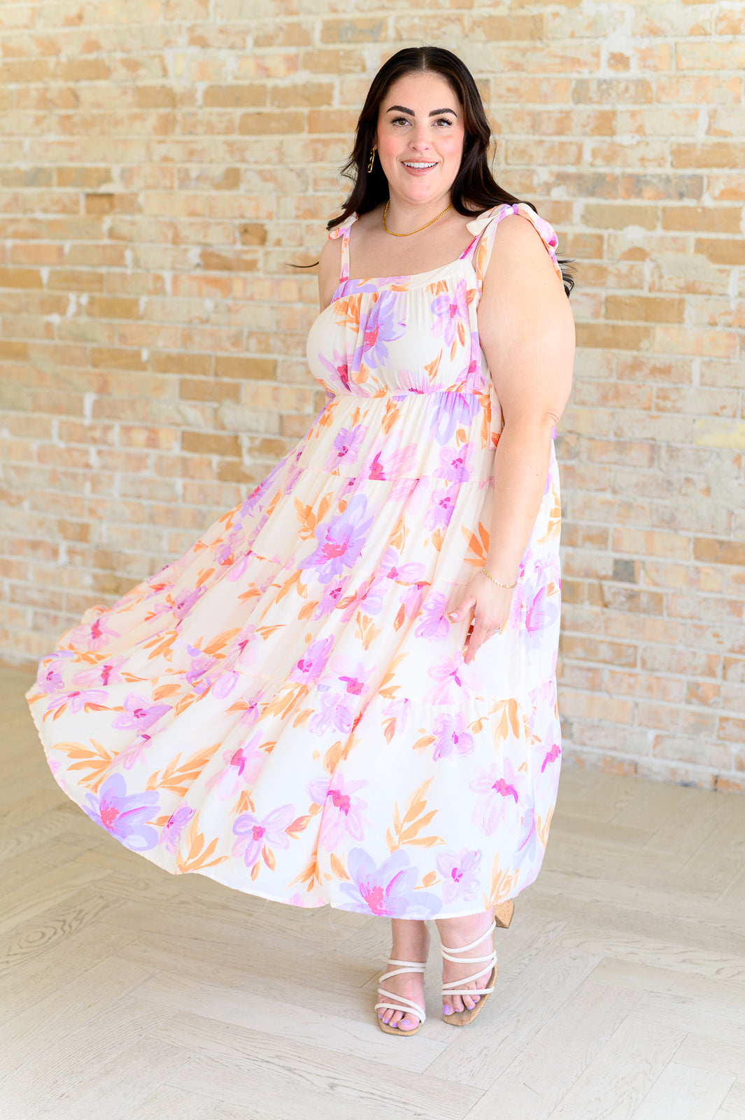 Smiling woman wearing pastel floral midi dress with shoulder ties and sandals against light brick wall background