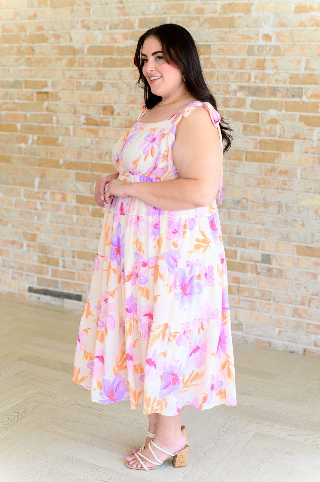 Smiling plus-size woman wearing pastel petals floral midi dress with shoulder ties and block heels by brick wall