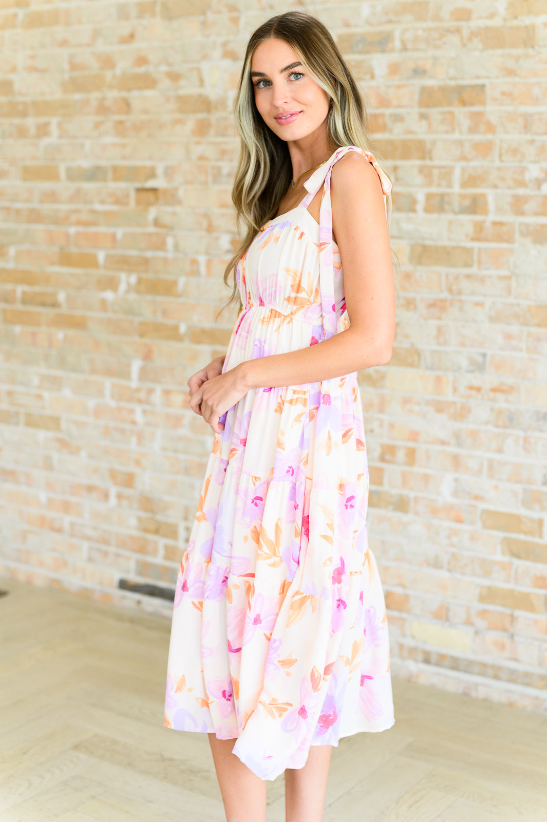 Woman in peach and lavender floral midi dress with tie shoulder straps standing against light brick wall