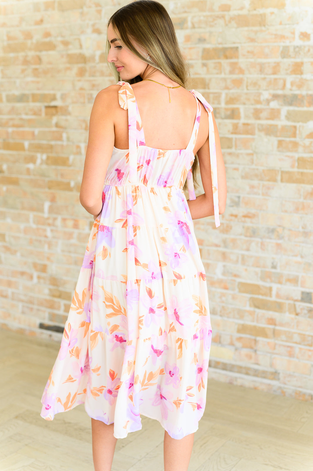 Woman wearing pastel peach floral midi dress with shoulder ties standing against light brick wall