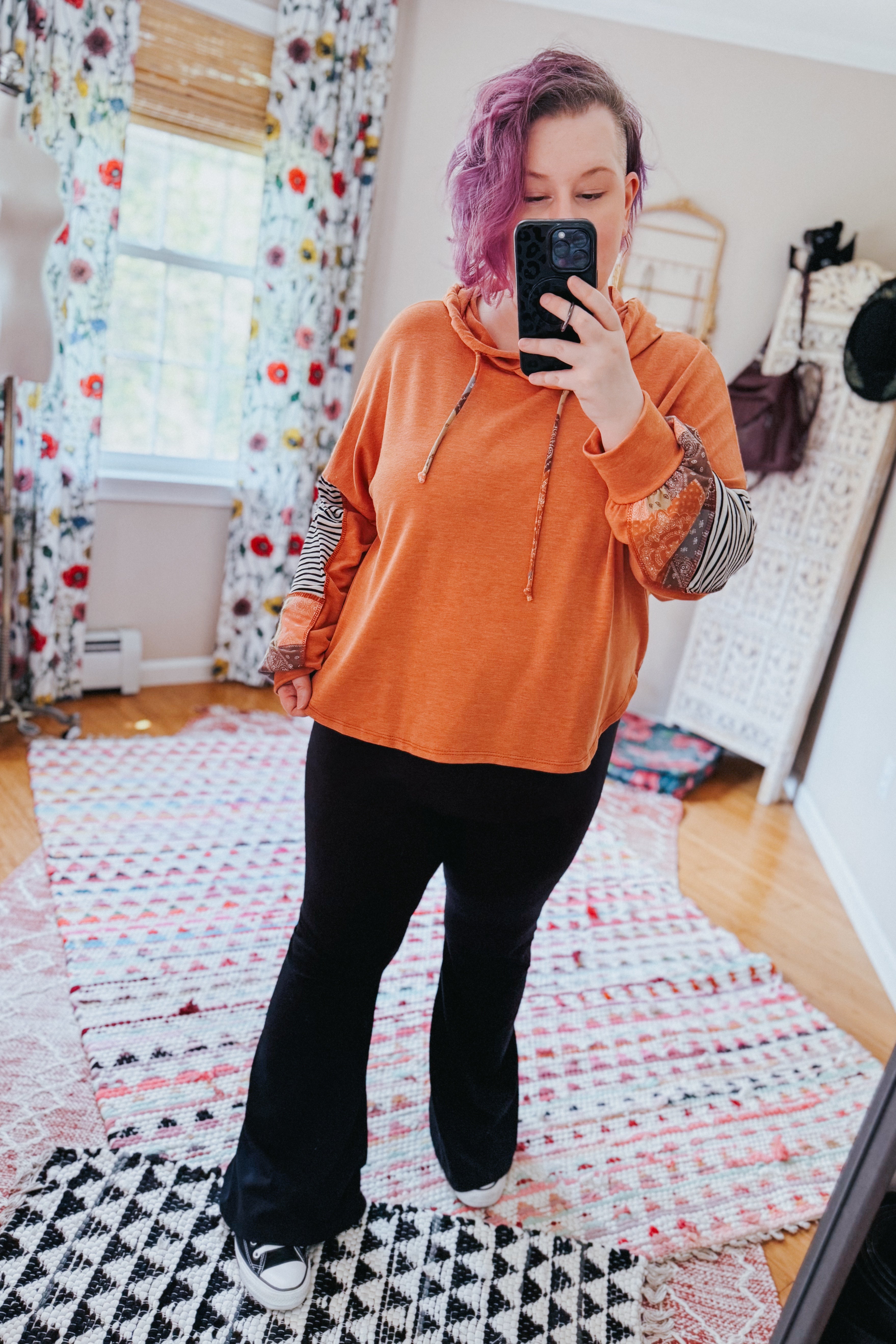 Woman with purple hair taking mirror selfie wearing rust hoodie with eclectic patterned sleeves and black pants indoors