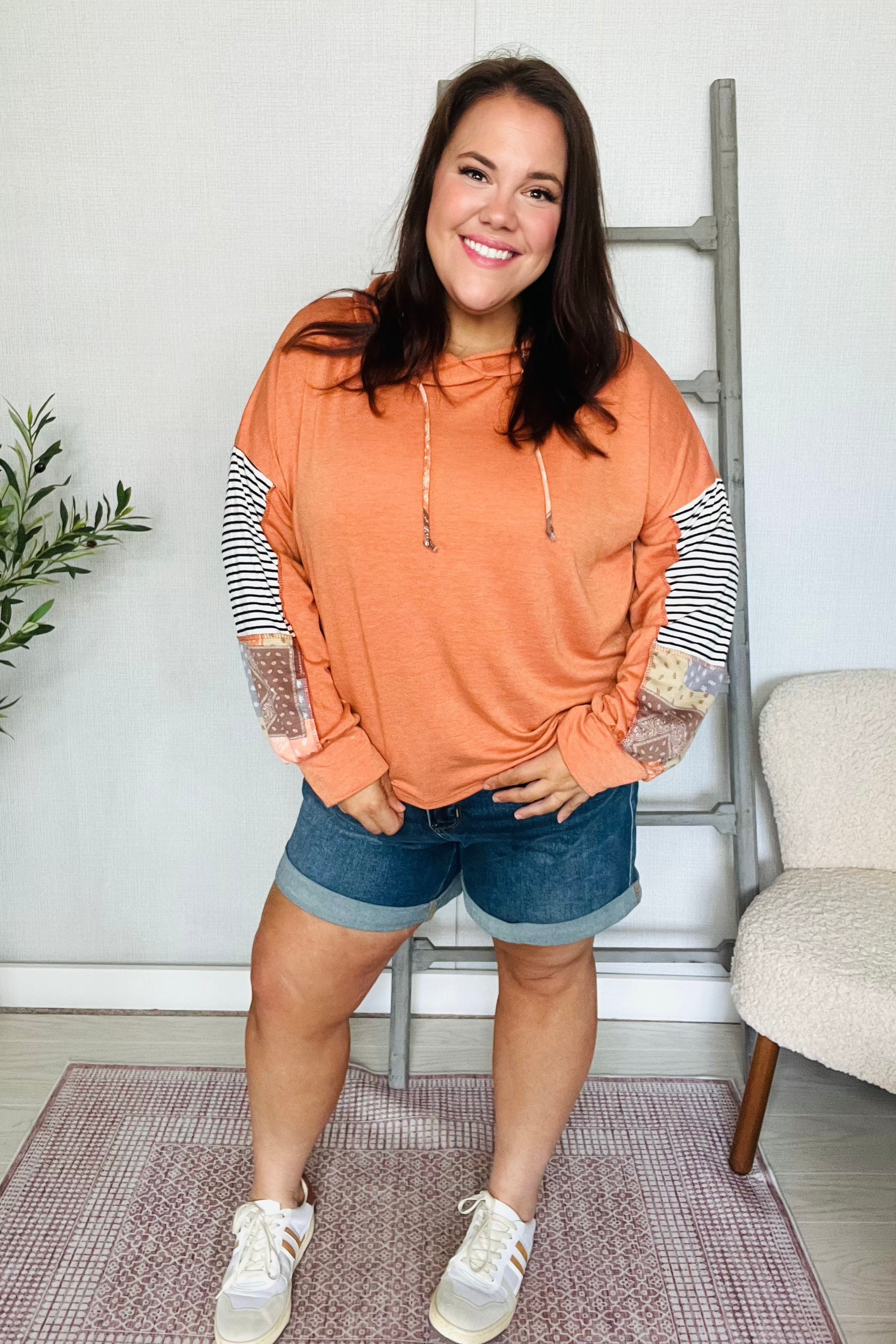 Smiling woman wearing rust hoodie with striped and patchwork sleeves paired with rolled denim shorts and sneakers indoors