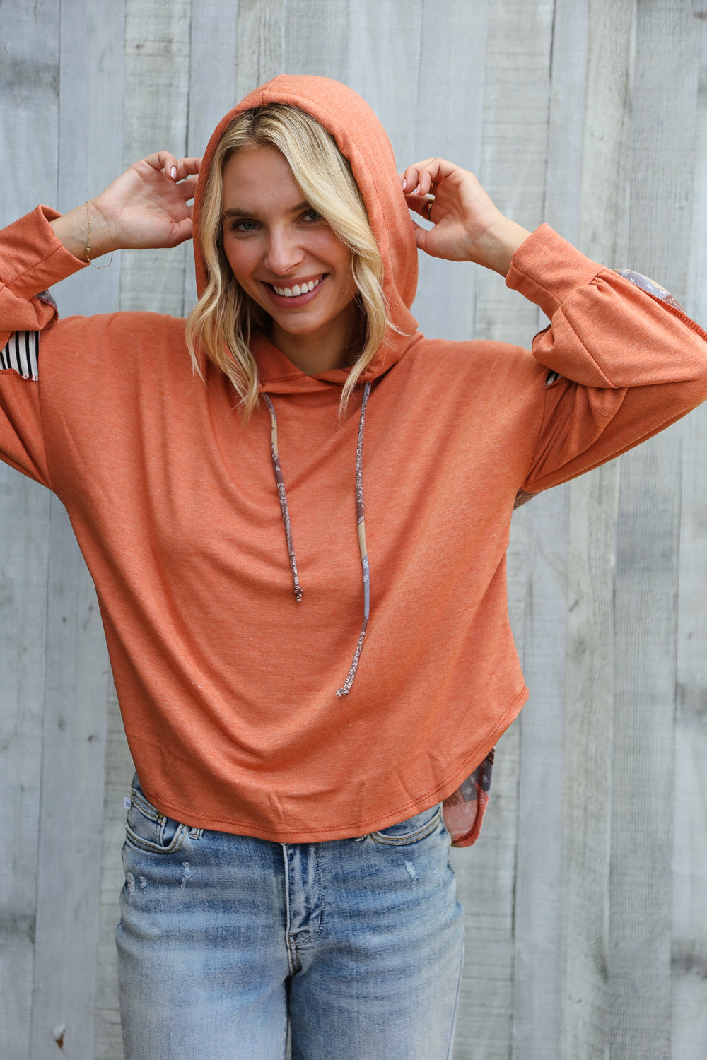 Smiling blonde woman wearing rust-colored patchwork French terry hoodie with hood up and casual light-wash jeans