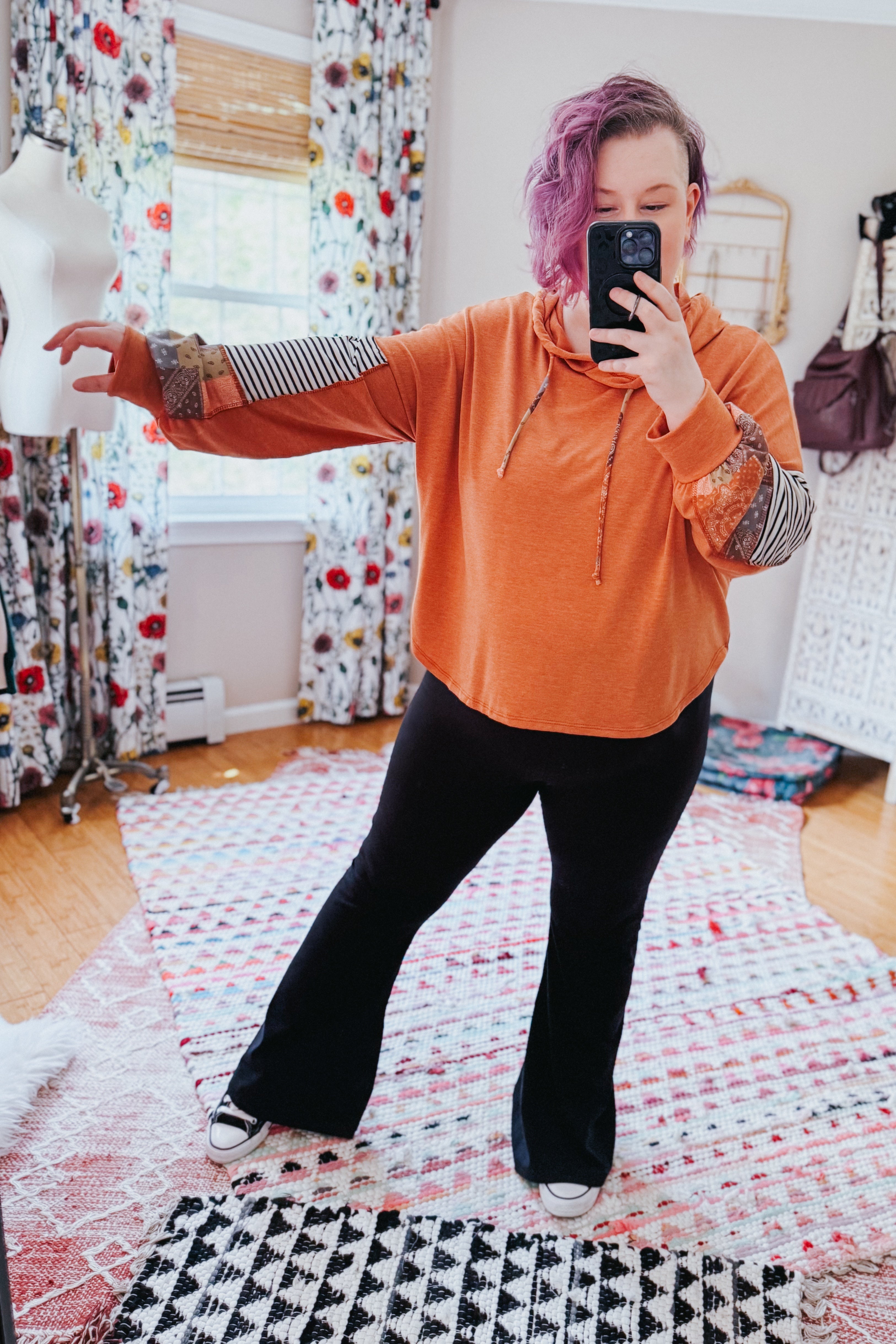Woman with purple hair taking selfie wearing rust patchwork hoodie with striped and patterned sleeves and black pants indoors