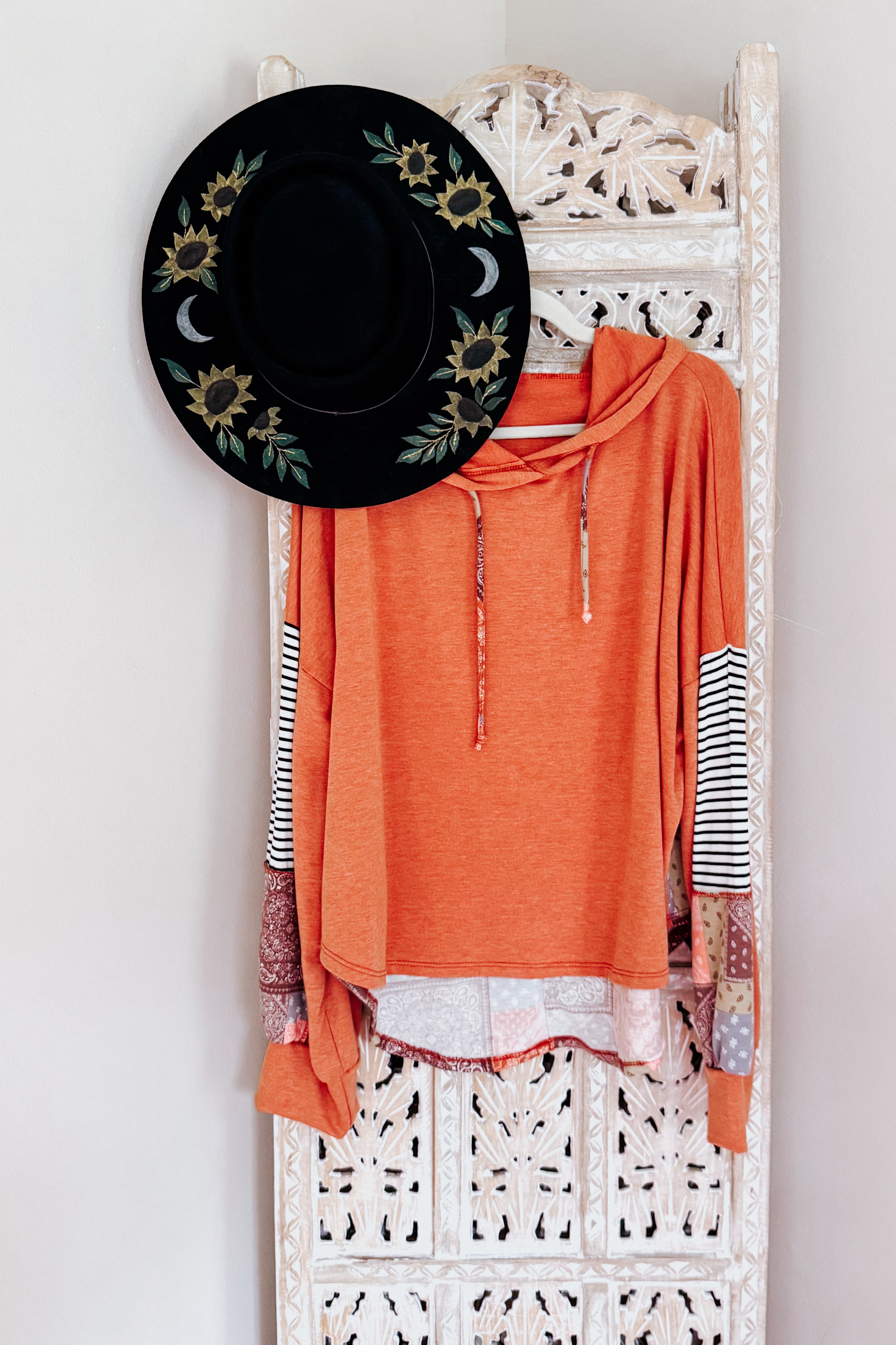 Rust-colored French terry hoodie with patchwork sleeves and striped accents hanging on carved wooden screen with black floral hat