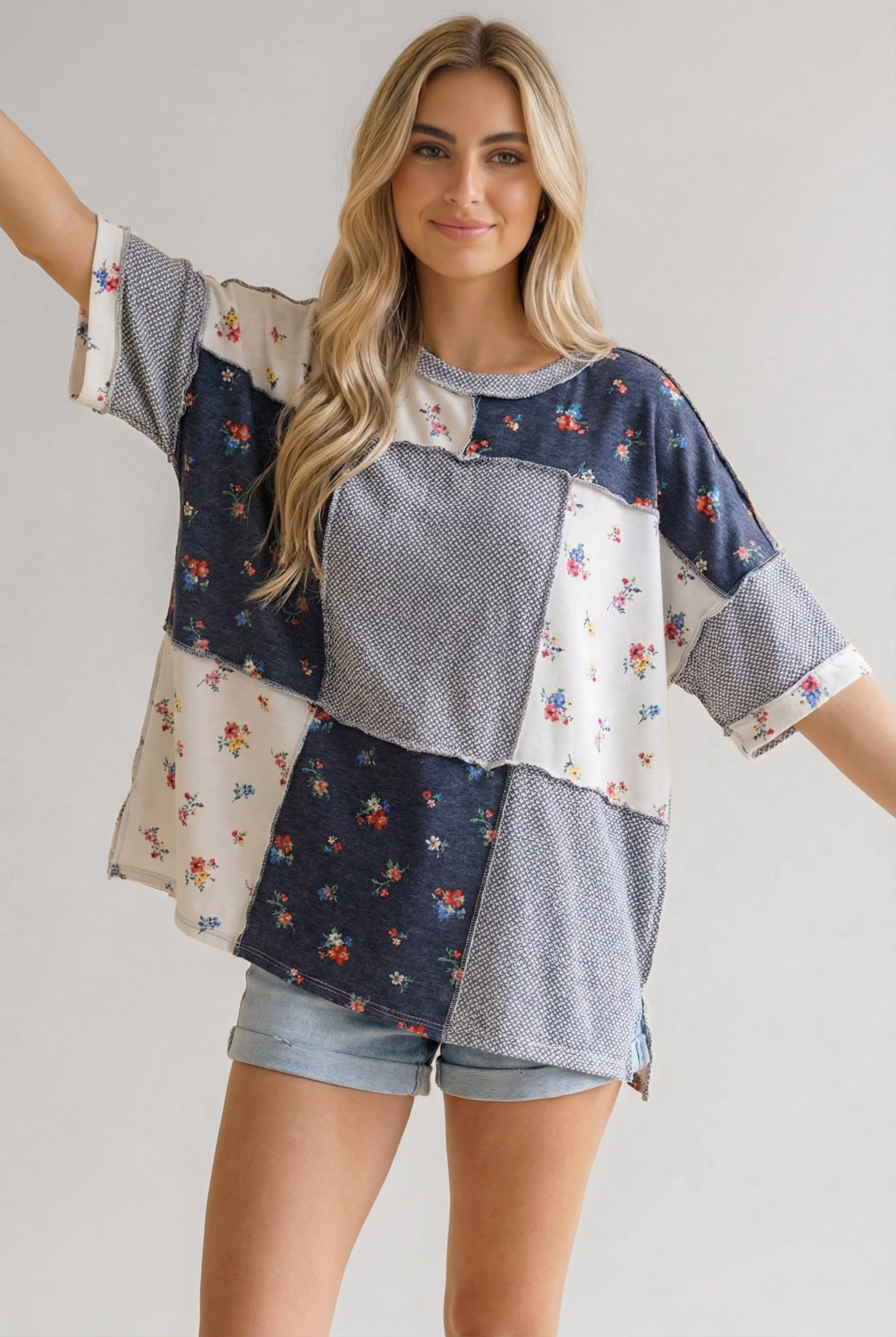 Woman wearing a loose blue patchwork top with small floral prints and textured knit panels paired with light denim shorts