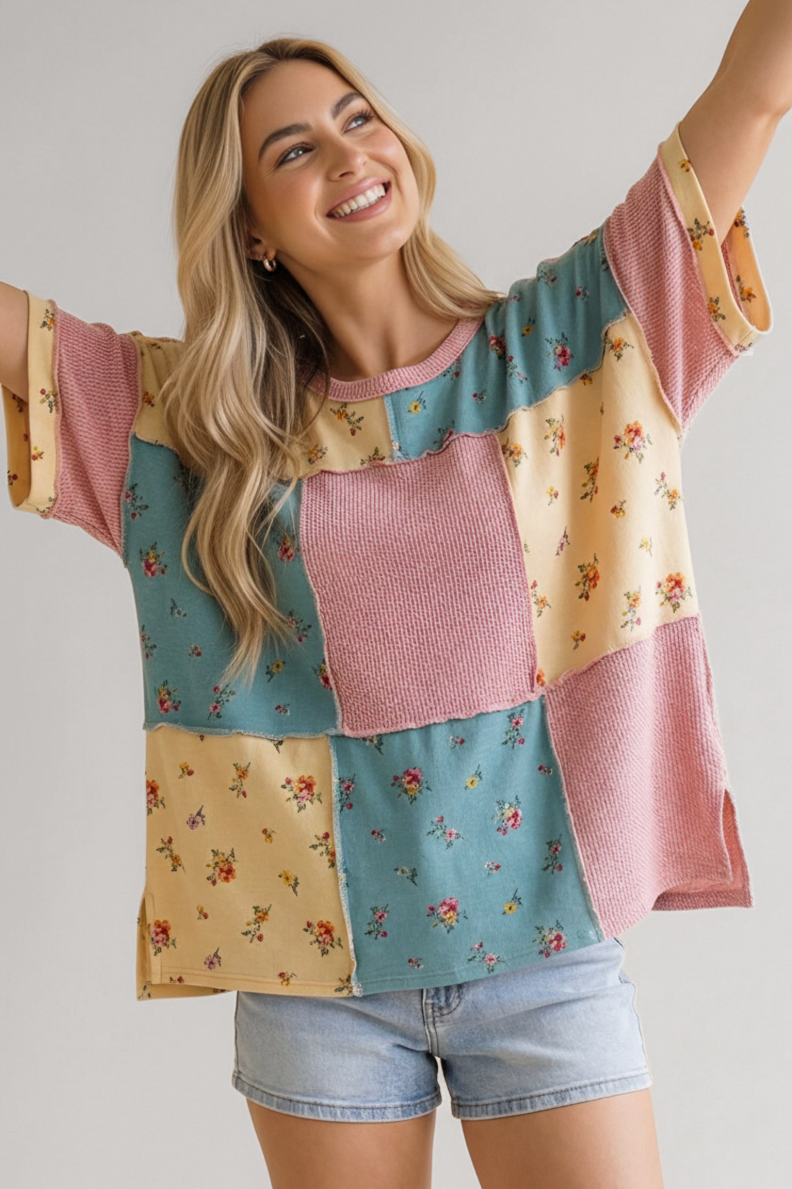 Smiling woman wearing a patchwork waffle knit top in pink, blue, and yellow with floral details and denim shorts indoors