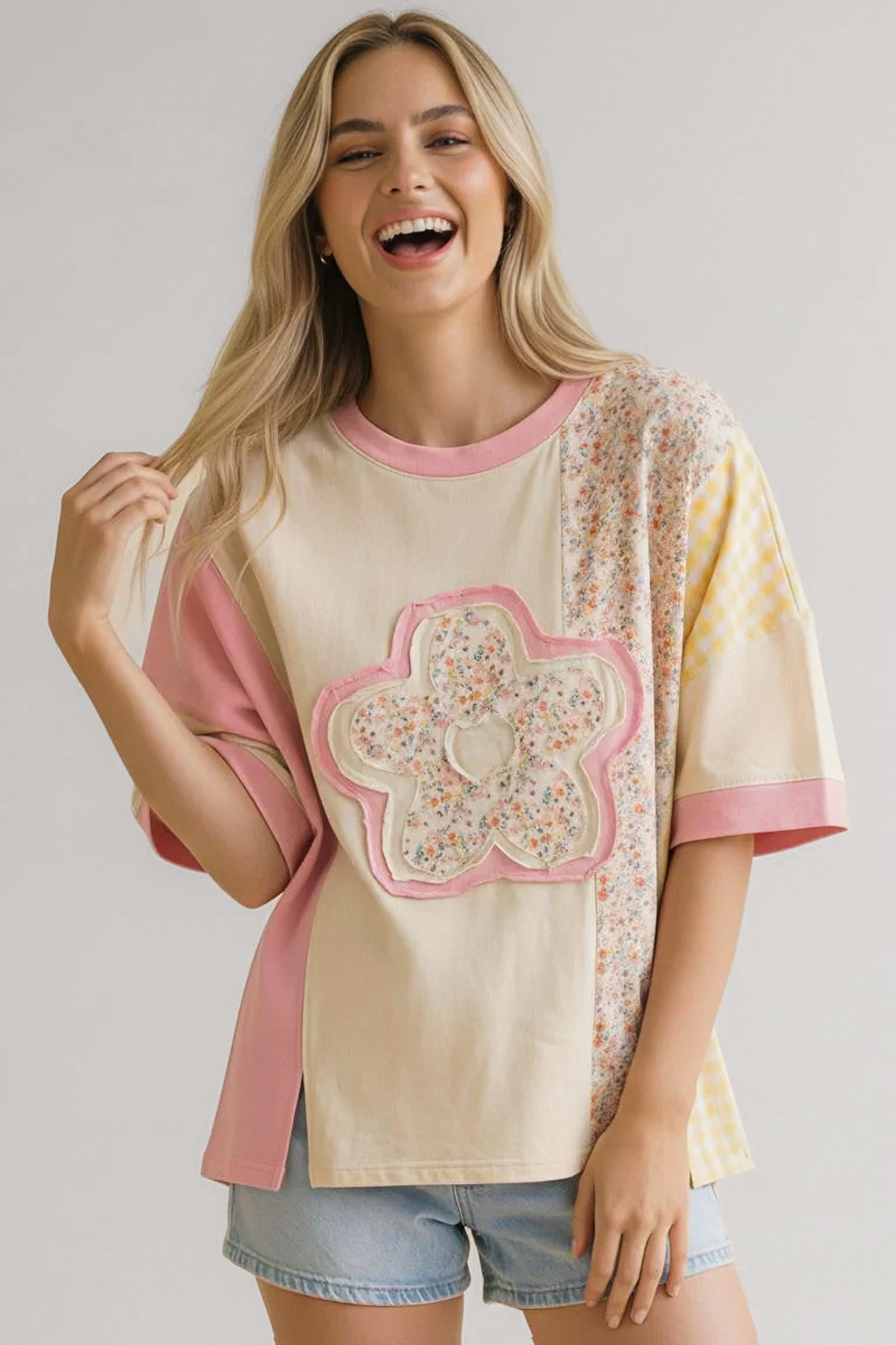 Smiling woman wearing relaxed pink floral patchwork tee with oversized flower applique and denim shorts indoors