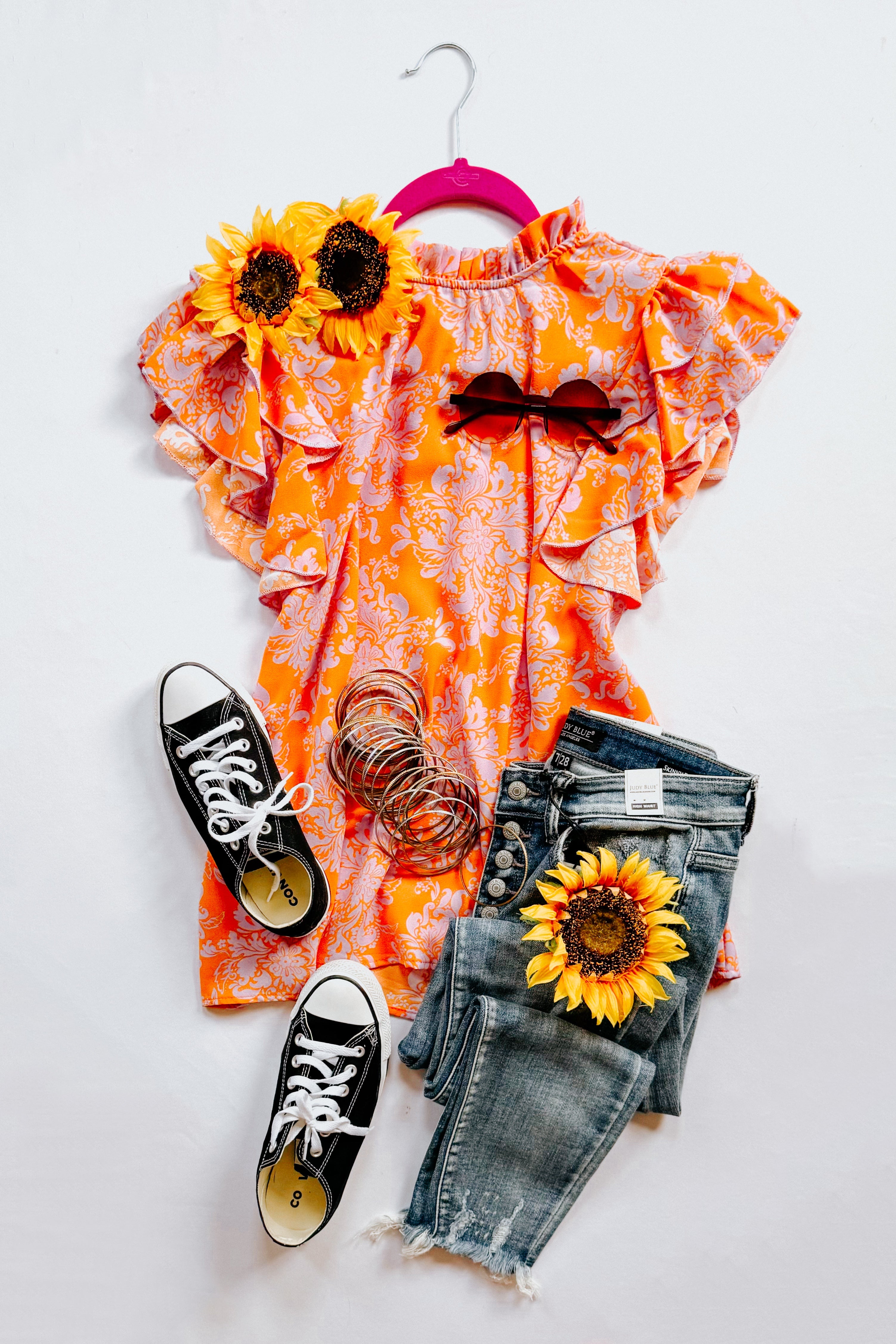 Orange paisley flutter sleeve top styled with denim jeans, black sneakers, sunglasses, bangles, and sunflowers on white background