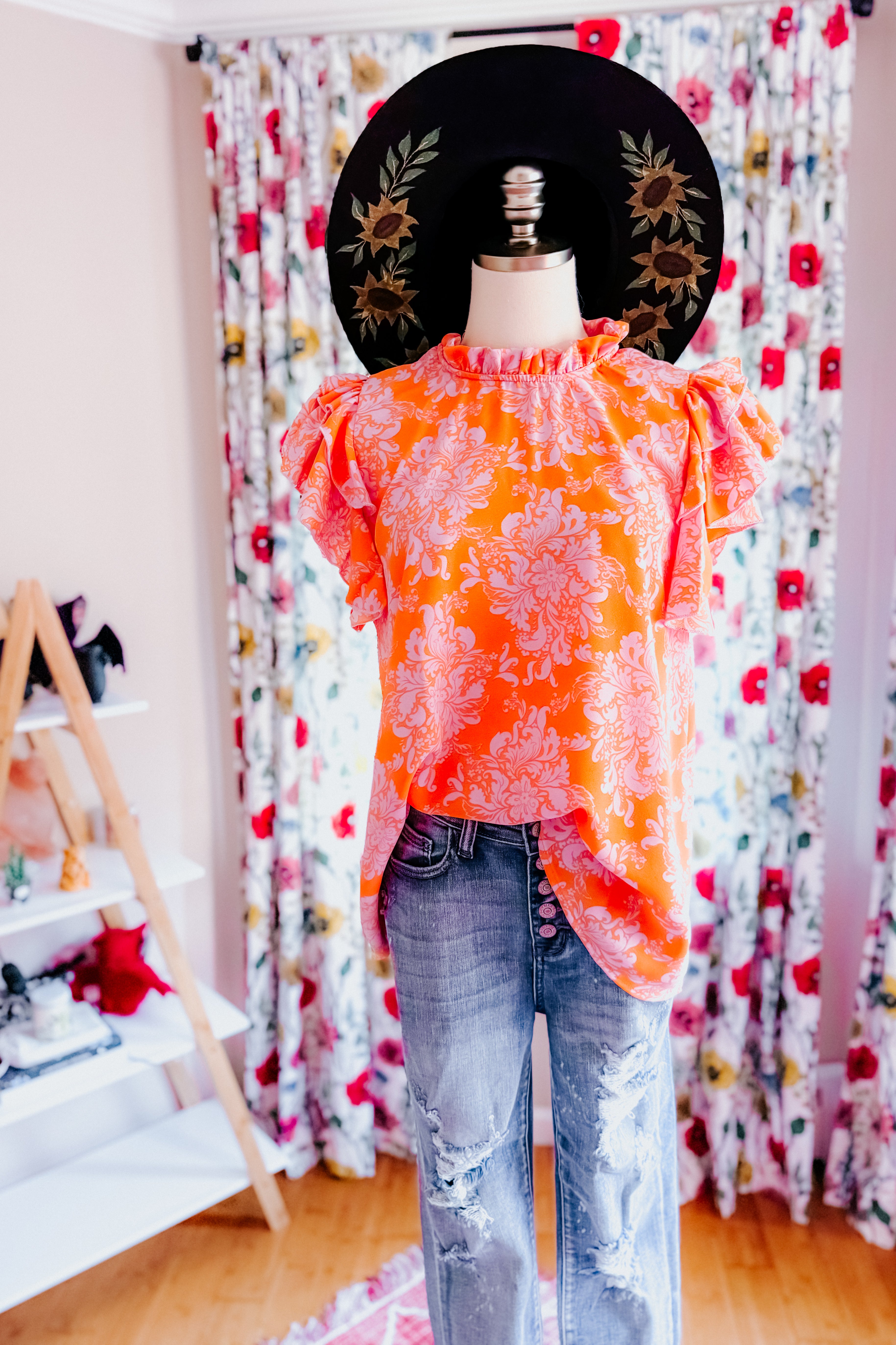 Orange paisley flutter sleeve top with mock neck styled with distressed denim jeans and black floral hat on mannequin