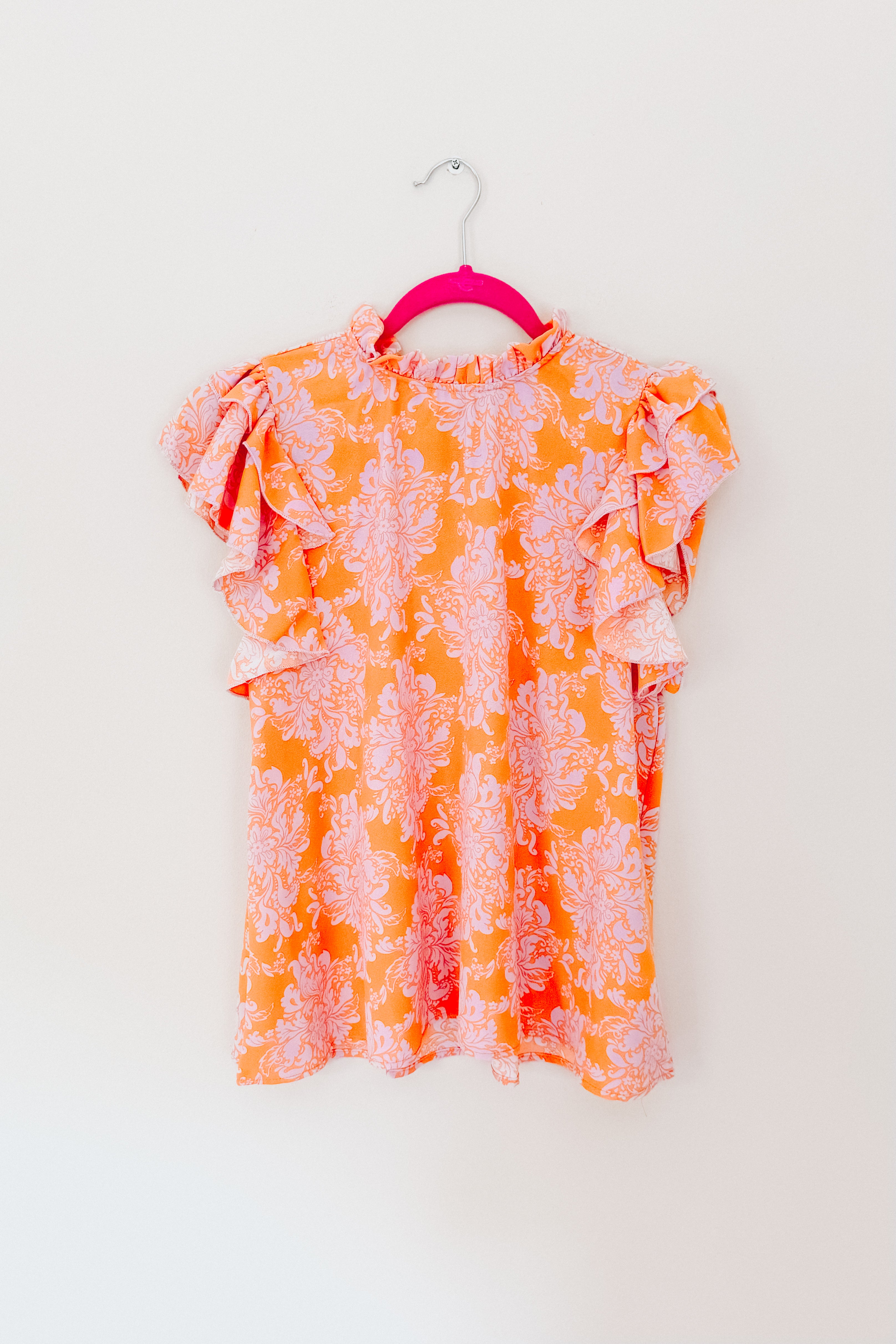 Orange paisley patterned flutter sleeve top with mock neck hanging on bright pink hanger against white background