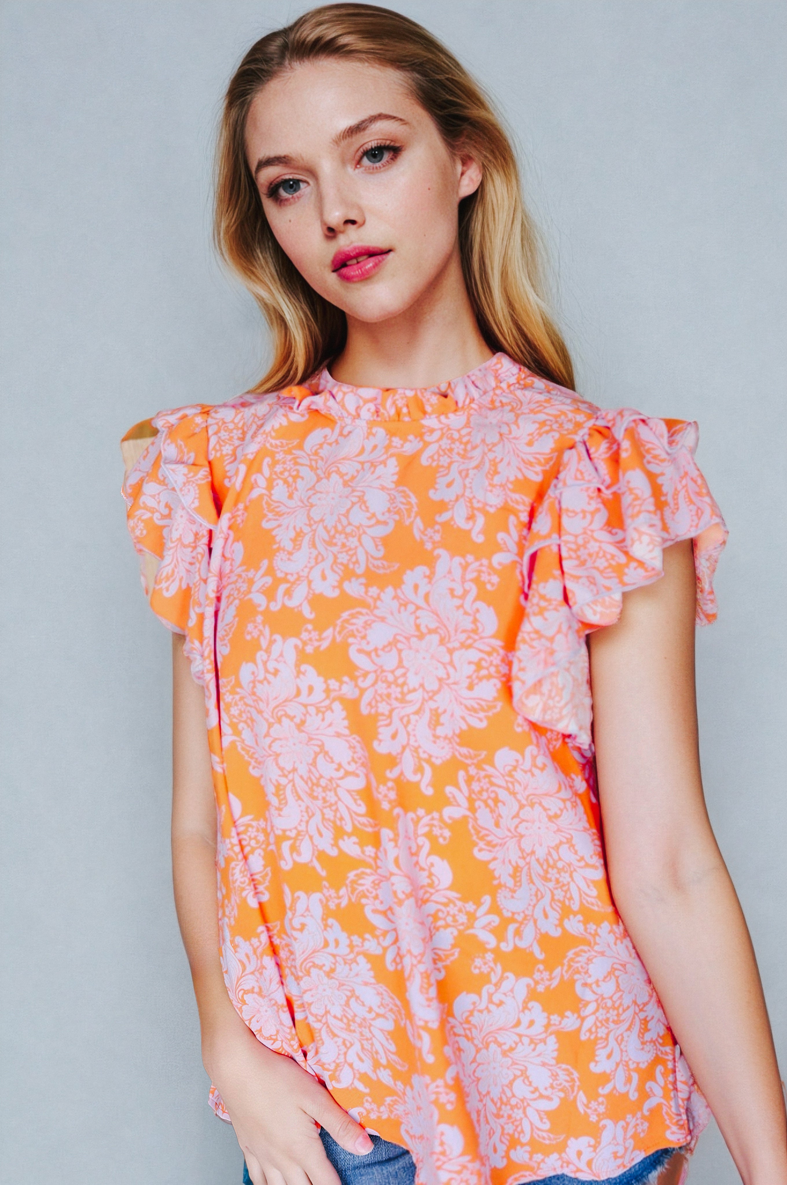Woman wearing orange paisley patterned top with ruffled flutter sleeves and mock neck against light background