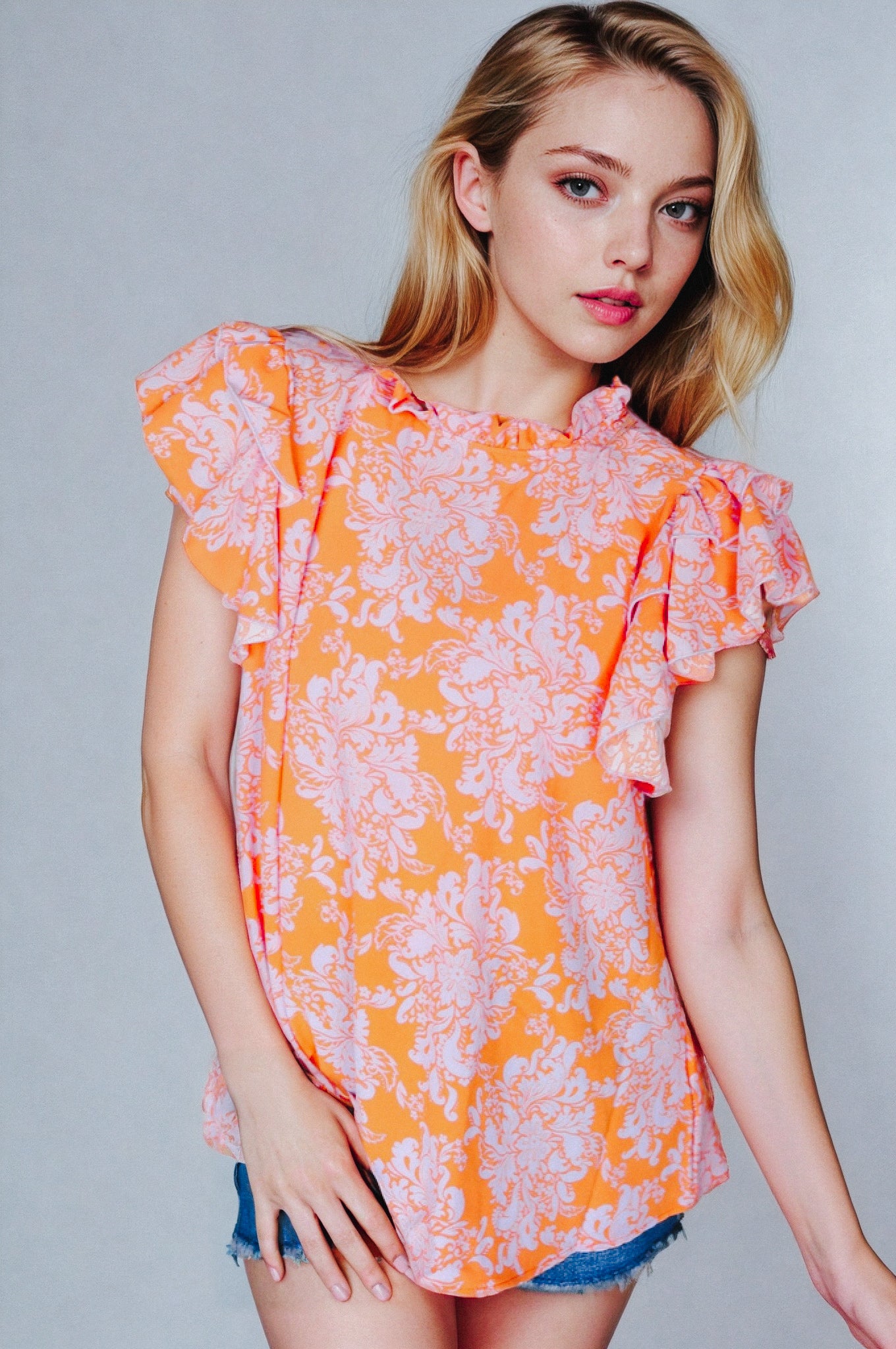 Blonde woman in orange paisley top with ruffled flutter sleeves and mock neck posing against a gray backdrop