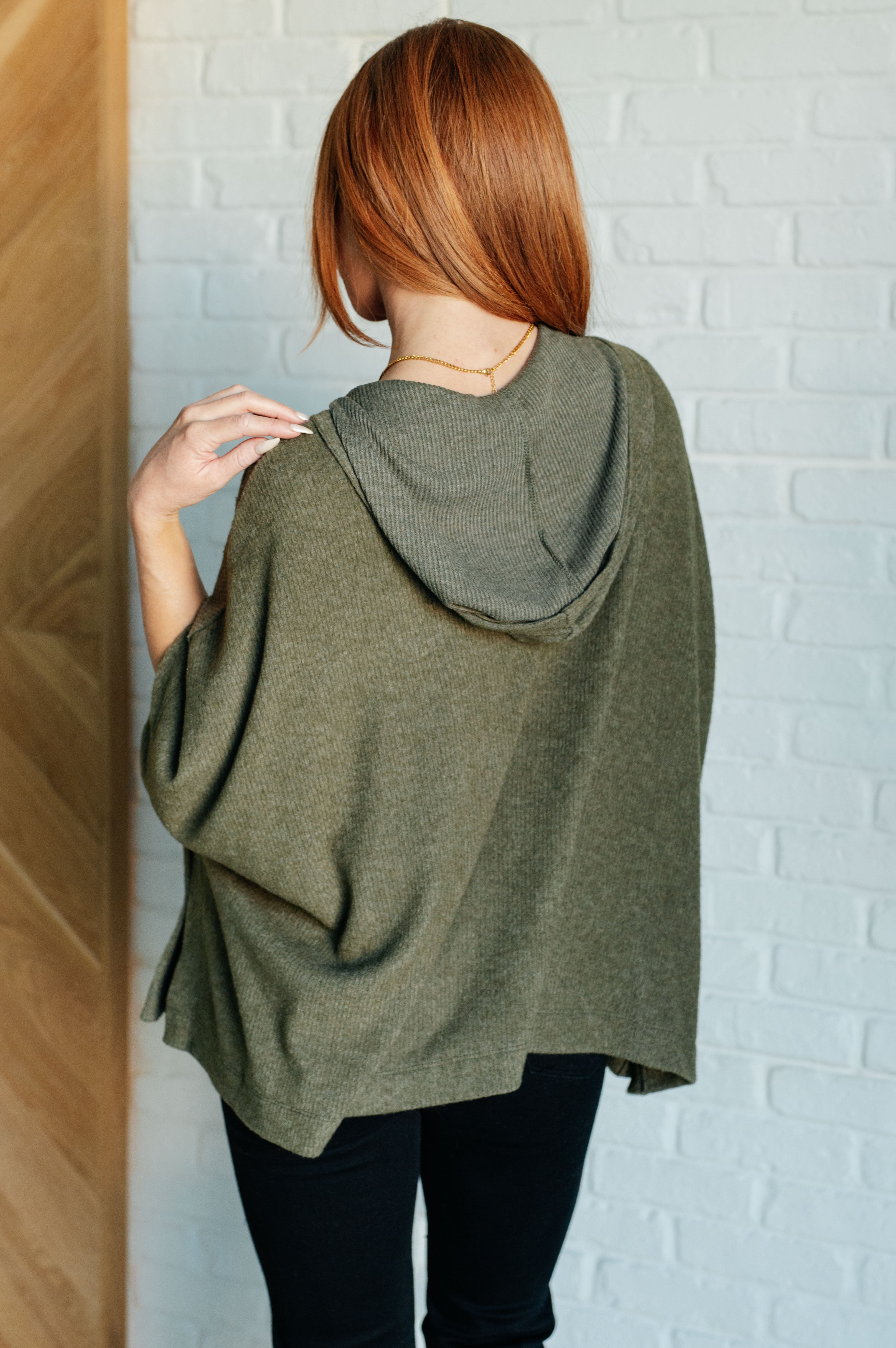 Woman with red hair wearing an olive hooded poncho with relaxed fit styled with black pants indoors