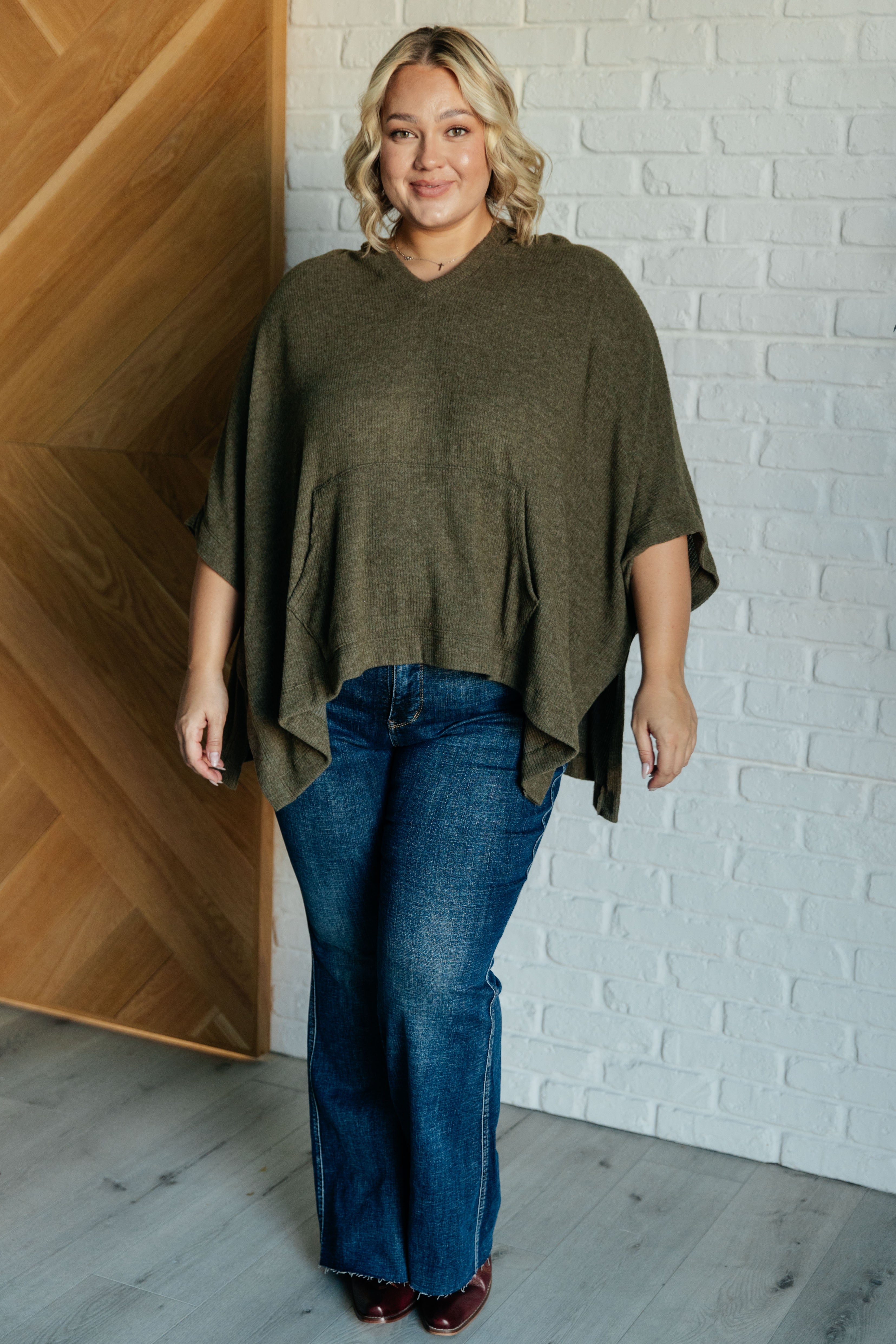 Smiling plus-size woman wearing an olive hooded poncho styled with blue jeans indoors against white brick wall