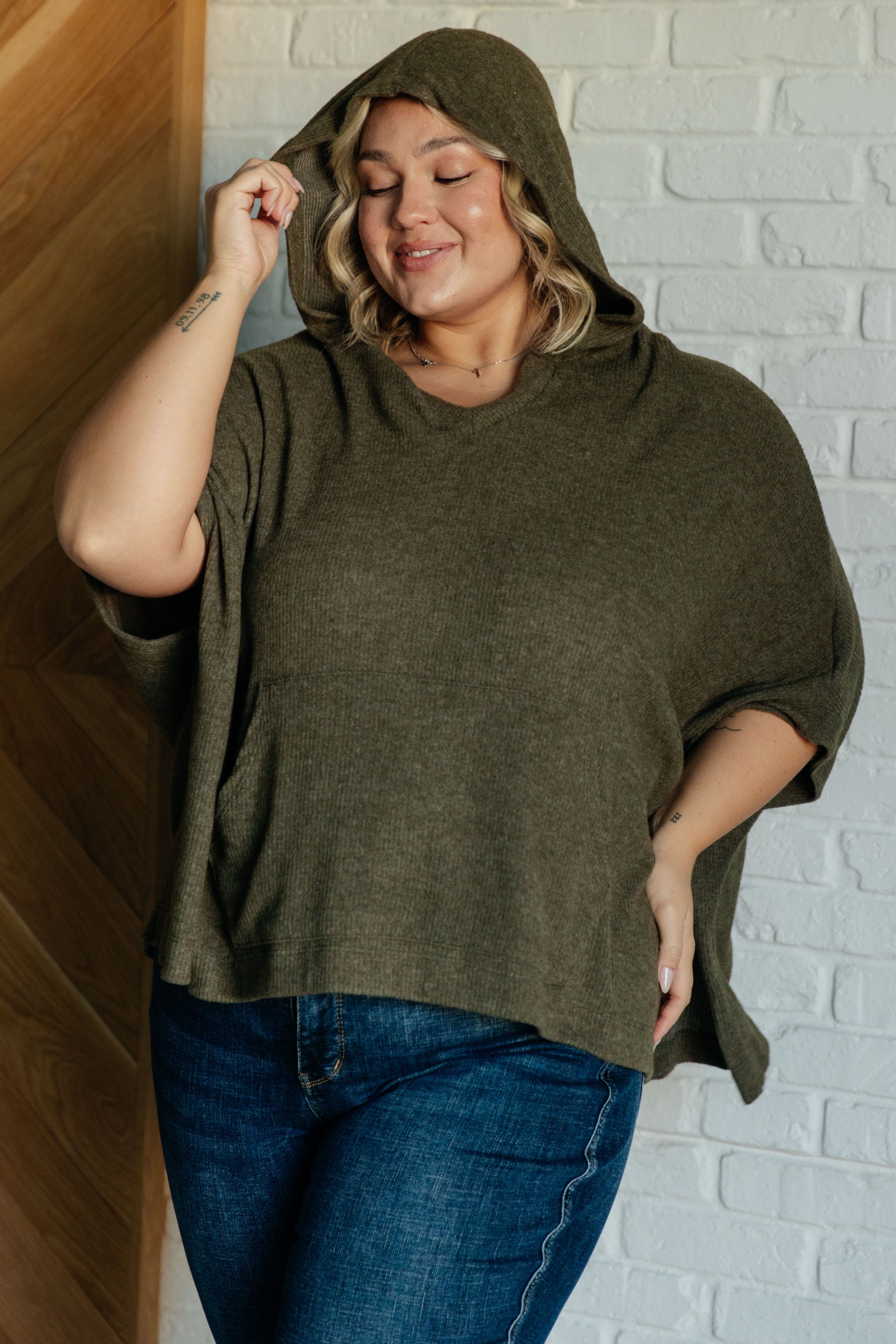 Plus-size woman wearing an olive hooded poncho with relaxed fit styled with blue jeans in indoor setting