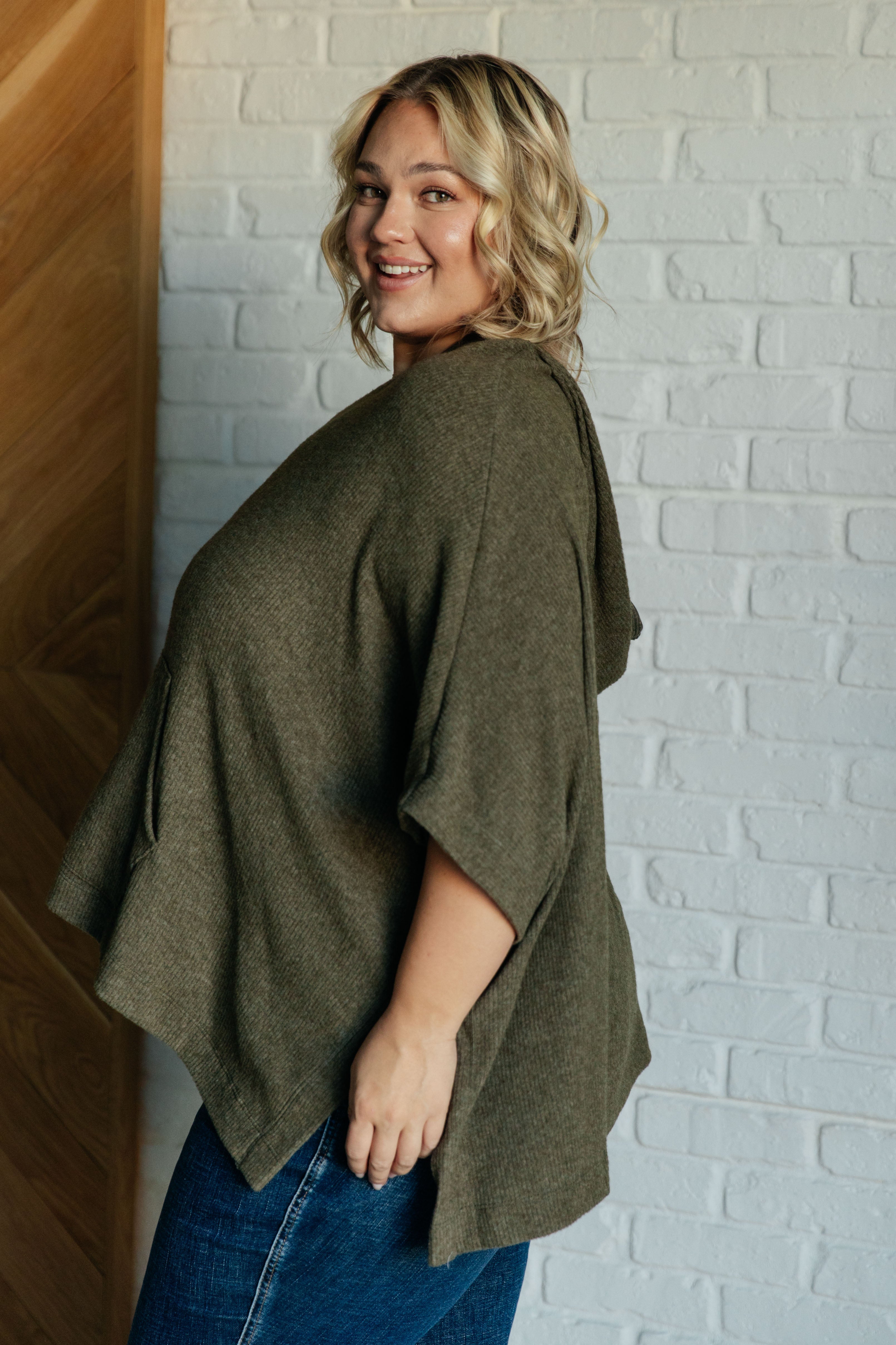 Plus-size woman wearing an olive knit hooded poncho with side pockets, styled with blue denim in casual indoor setting