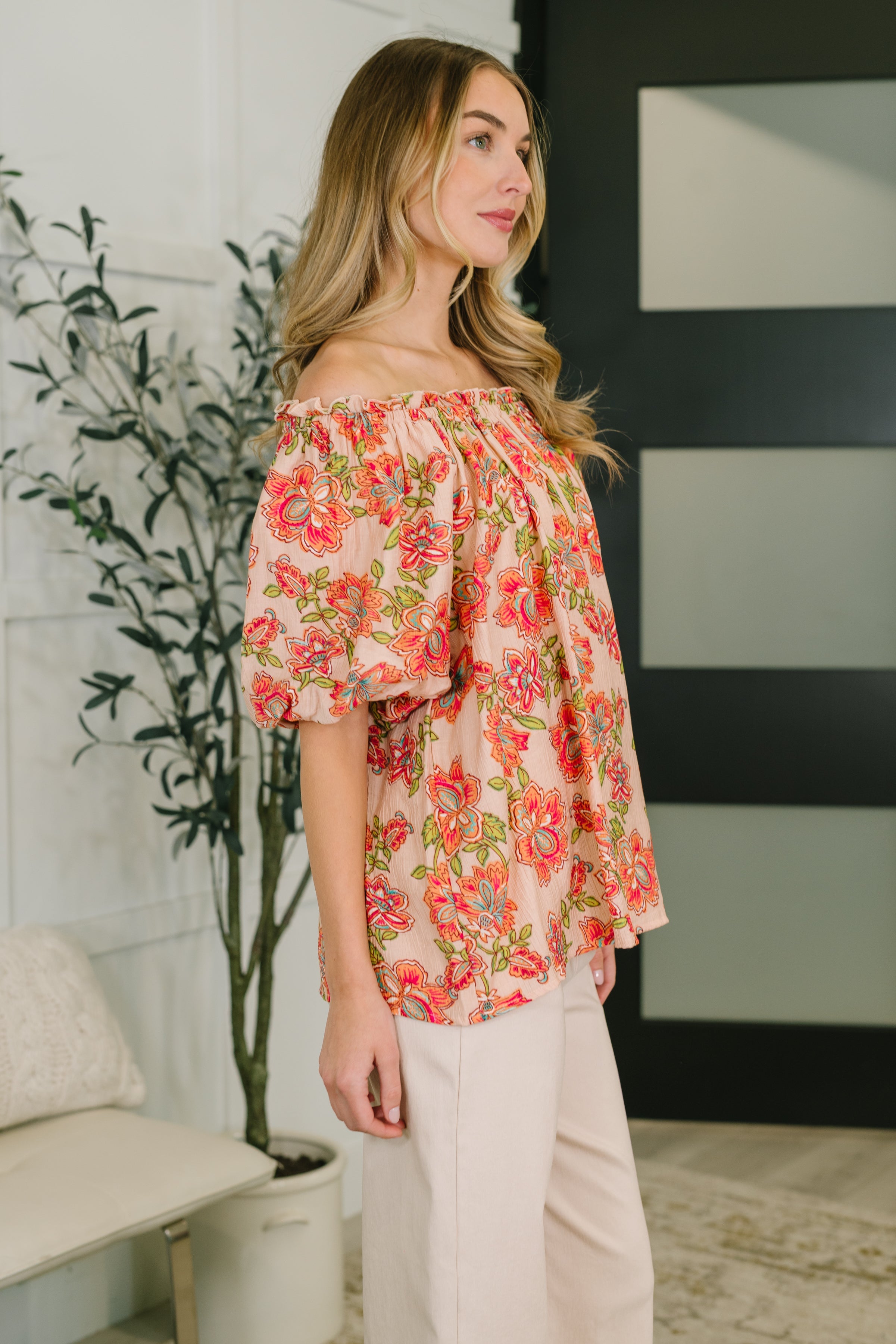Side view of woman wearing orange floral off-shoulder blouse with puff sleeves paired with light beige pants indoors
