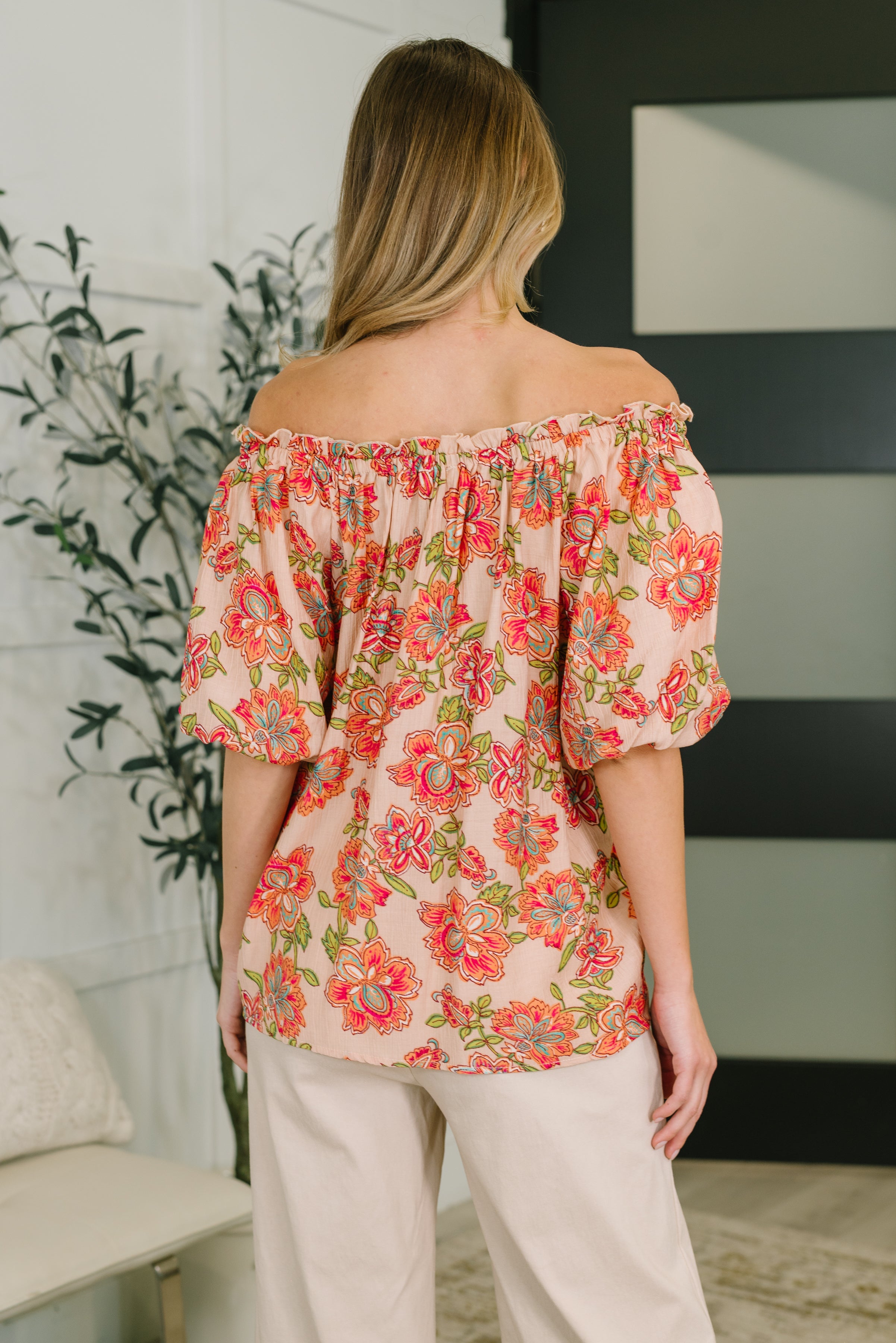 Back view of woman wearing a peach blouse with bright floral patterns and puff sleeves paired with light pants indoors