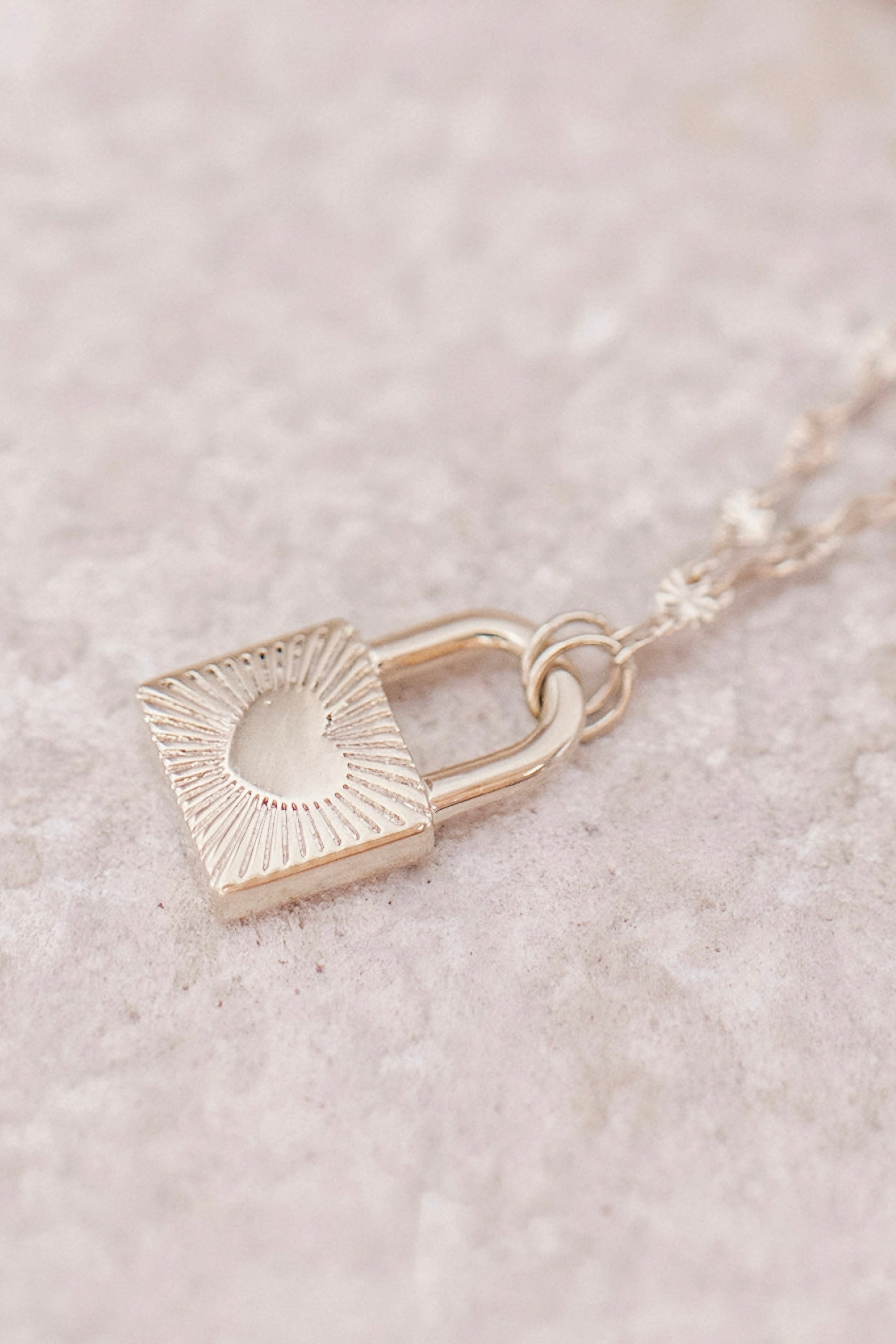 Delicate gold lock pendant with heart detail on fine chain resting on soft neutral surface in bold jewelry style