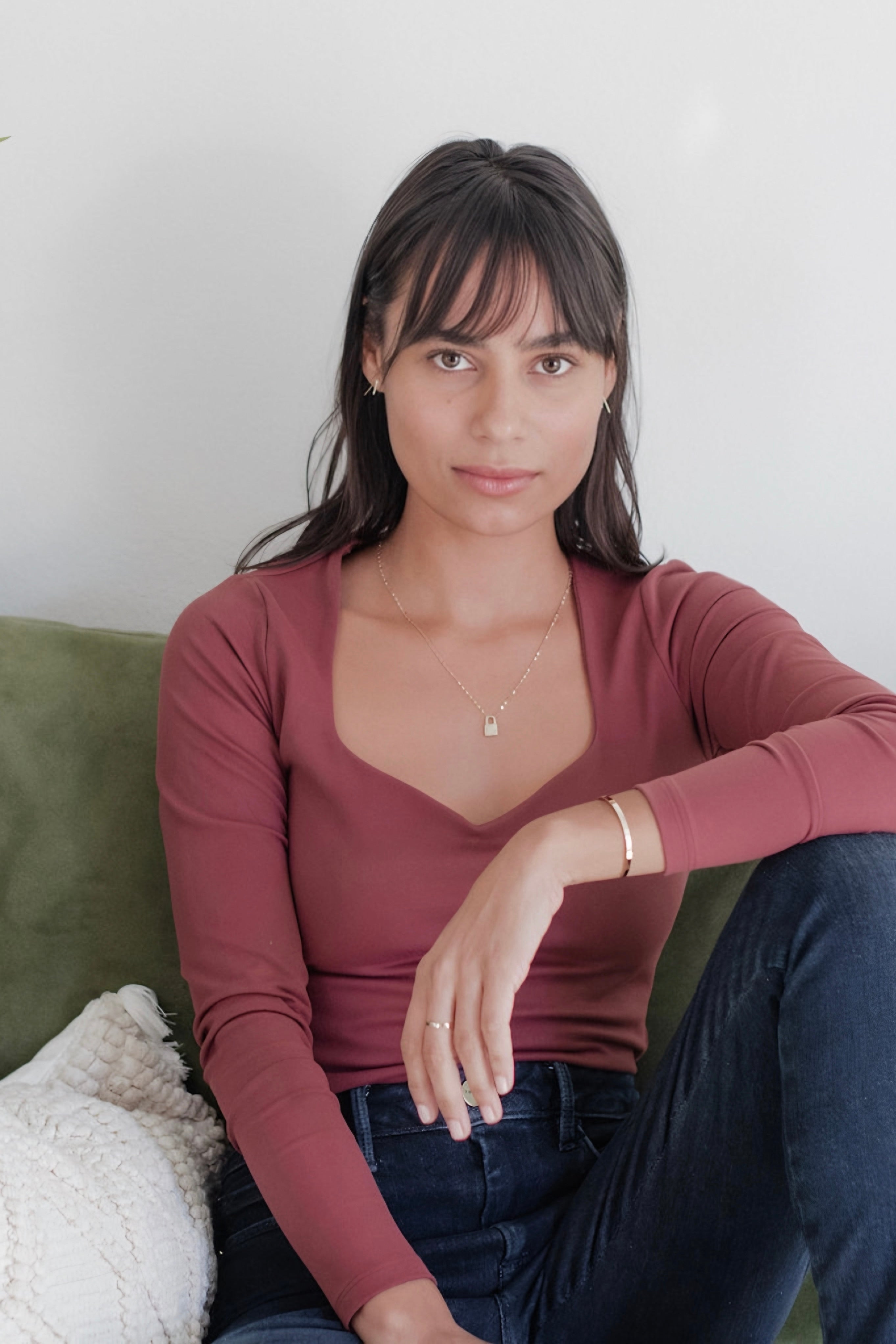 Woman seated wearing a gold lock pendant necklace with rose-colored square neckline top and dark jeans indoors