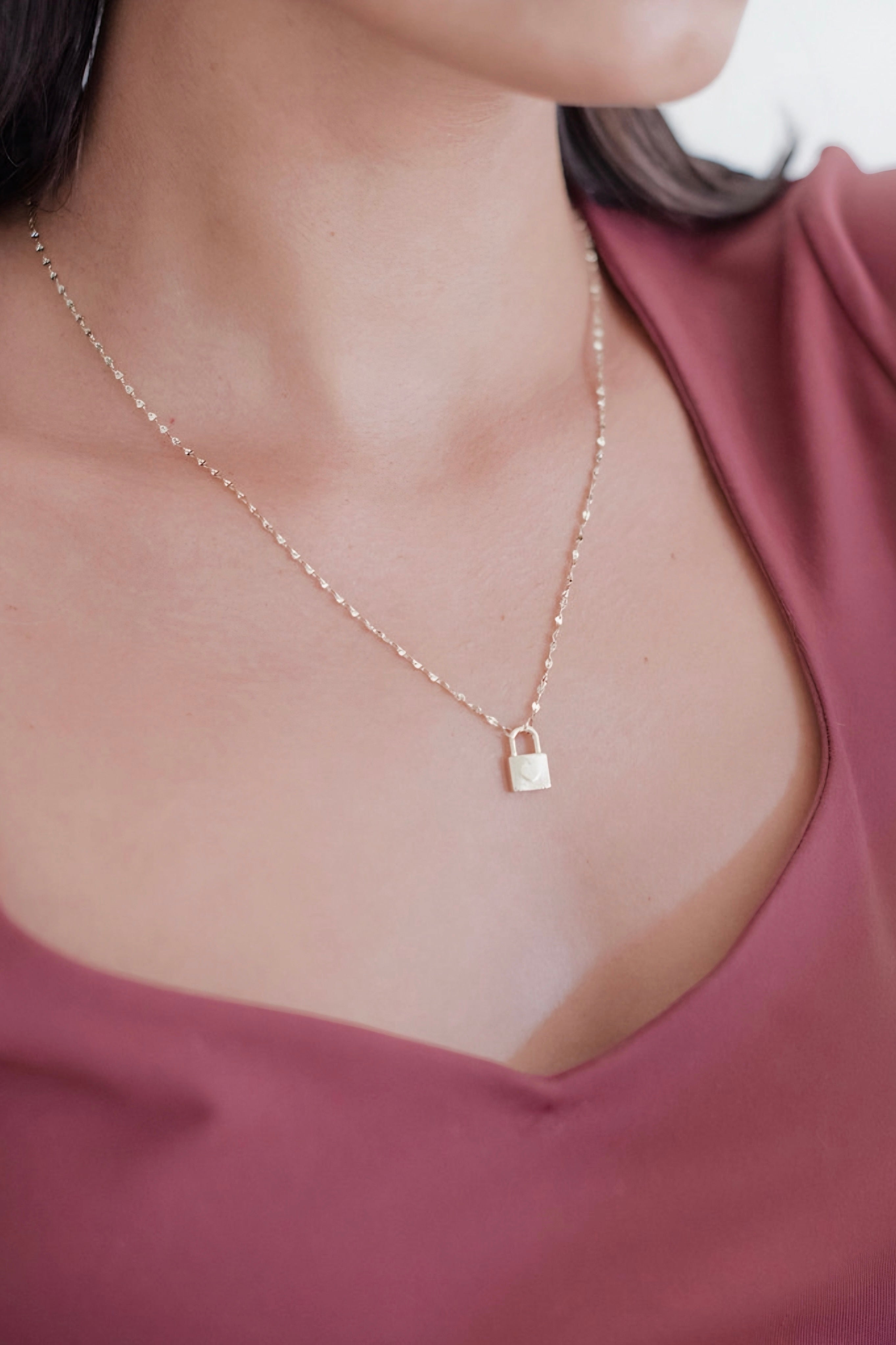 Close-up of gold lock charm necklace on delicate chain worn with rose-colored v-neck top in minimalist style